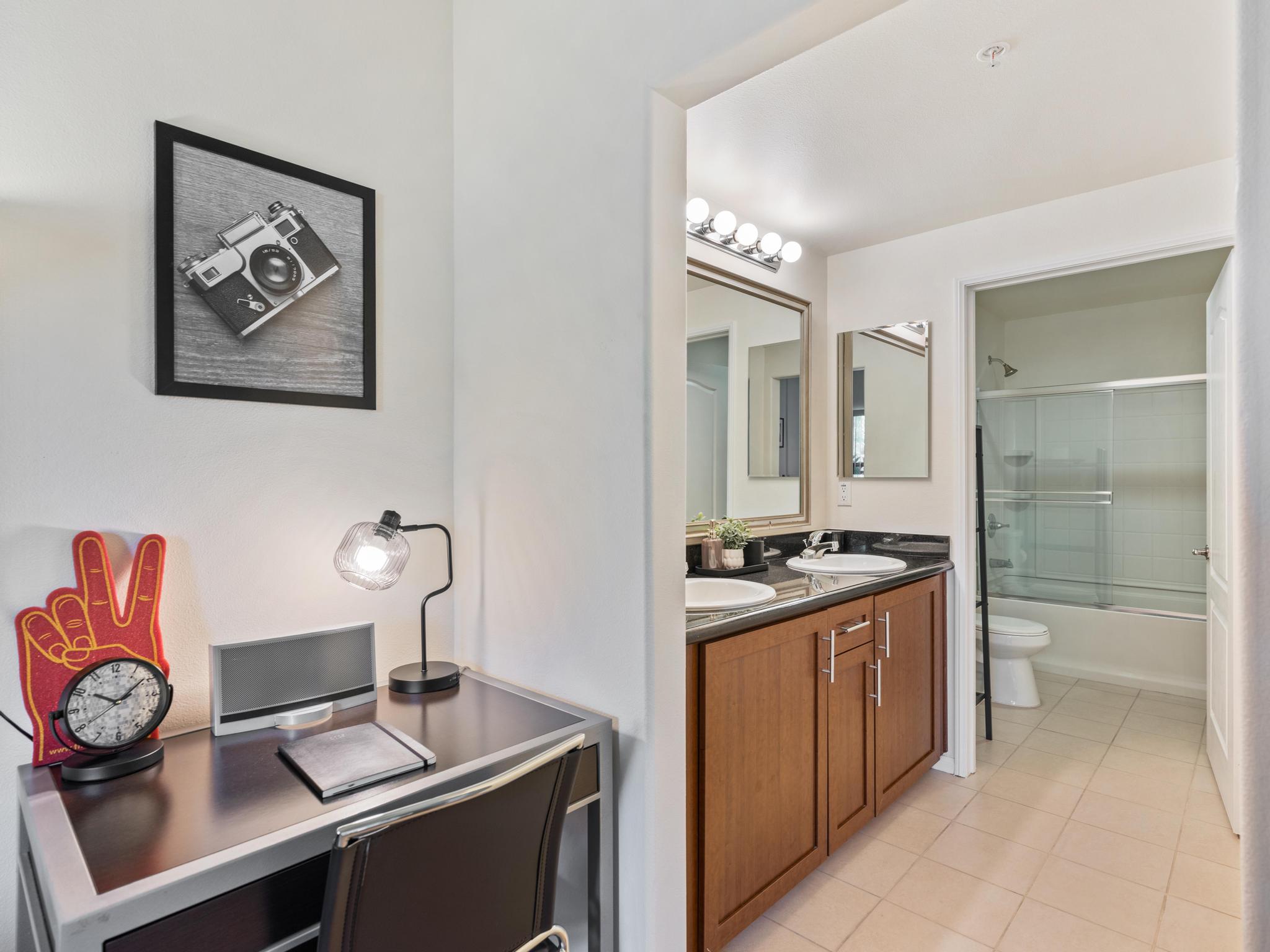 A bright and modern interior featuring a small workspace with a desk, lamp, and clock, adjacent to a clean bathroom with a large mirror, sink, and tub. A framed vintage camera photo decorates the wall, enhancing the contemporary aesthetic. The overall color scheme is light and inviting.