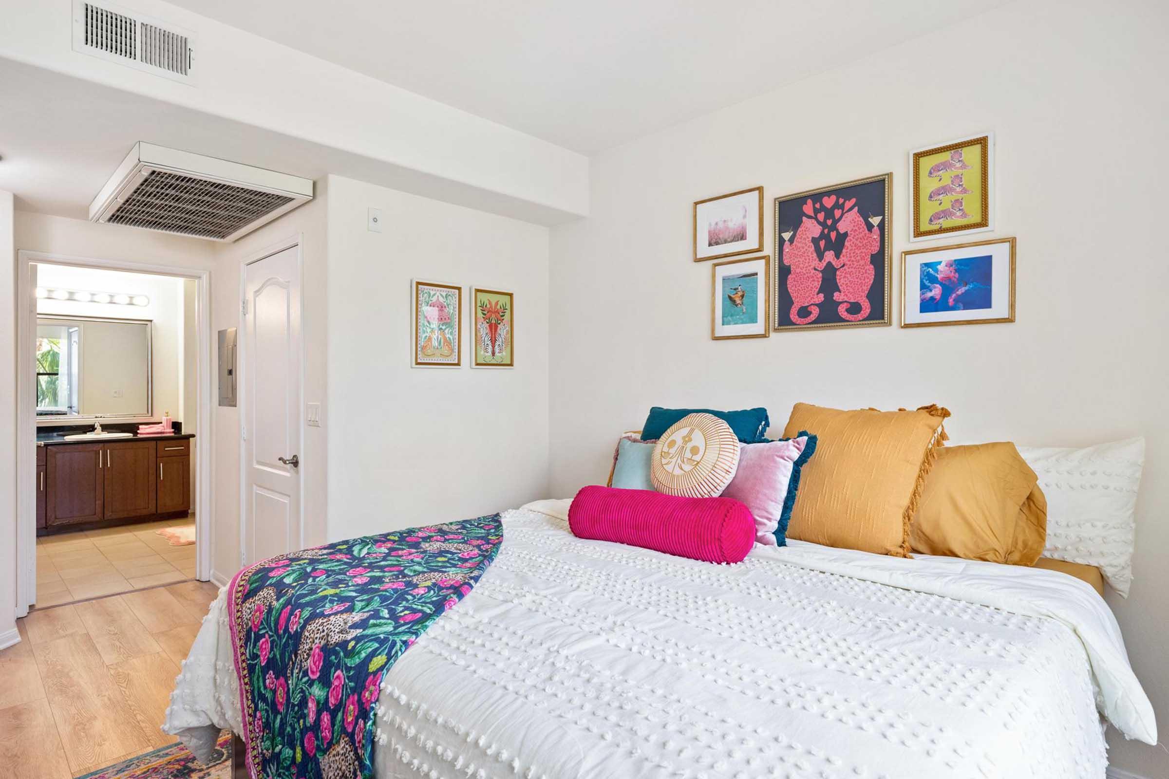 A cozy bedroom featuring a neatly made bed with colorful pillows and a patterned throw blanket. The wall above the bed is decorated with framed artwork. A doorway in the background leads to a bathroom, visible through an open door. The room has a bright and inviting ambiance.
