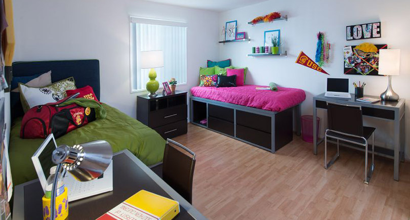 A cozy, modern bedroom setup featuring two beds with colorful bedding, a desk with a chair, and decorative shelves. The room has light wooden flooring and ample natural light, with decorative items and college-themed decor enhancing the lively atmosphere.