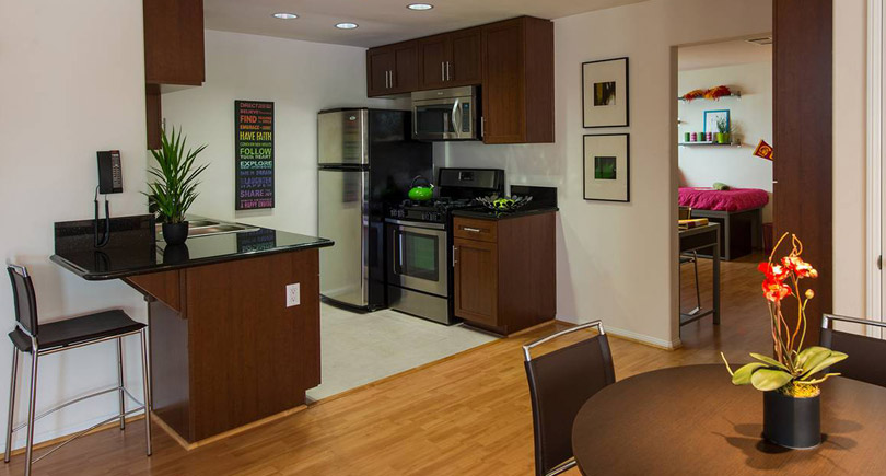 A modern kitchen featuring dark wood cabinets and stainless steel appliances. There's a black countertop with a small plant, bar stools nearby, and a dining table with a plant centerpiece. A doorway leads to another room with a pink bed visible in the background. The space is well-lit and contemporary in design.