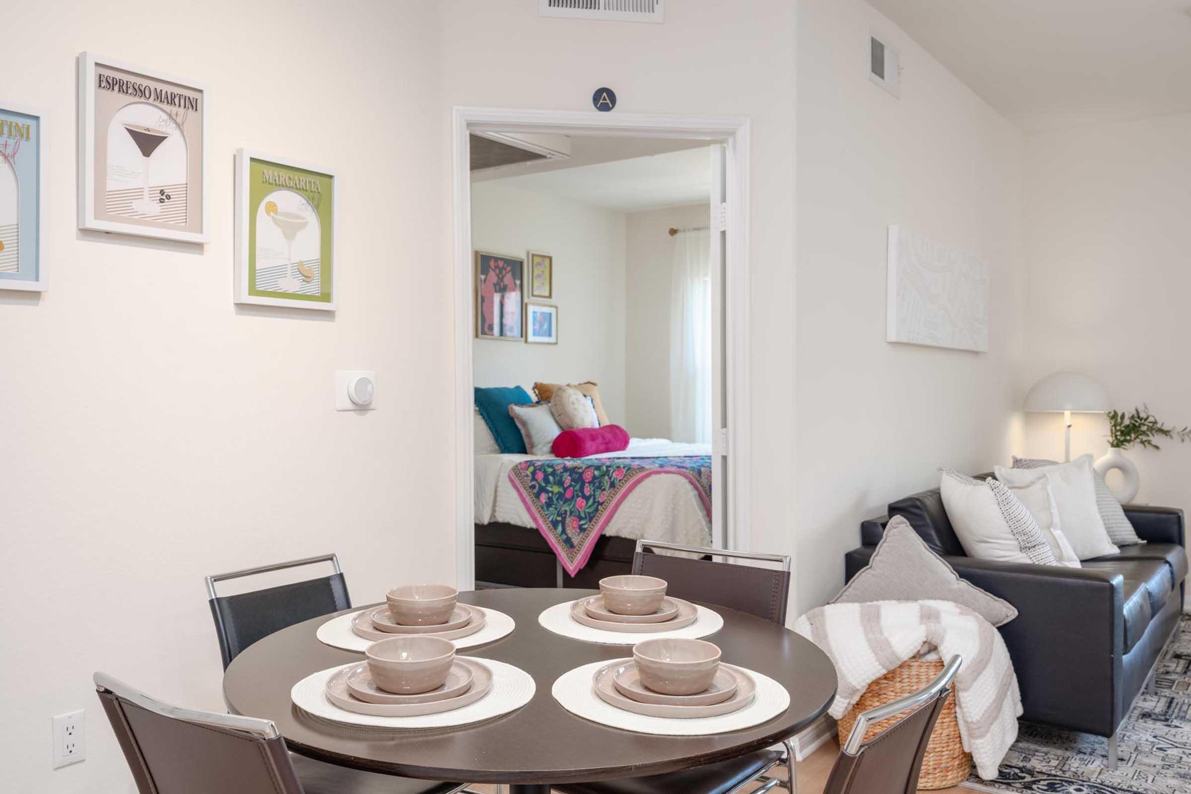 A modern living space featuring a round dining table set for four with beige dishes, a cozy sofa with decorative pillows and a throw blanket, and artwork framed on the wall. An open doorway reveals a bedroom with a bed dressed in colorful linens and additional decor, enhancing the inviting atmosphere of the room.