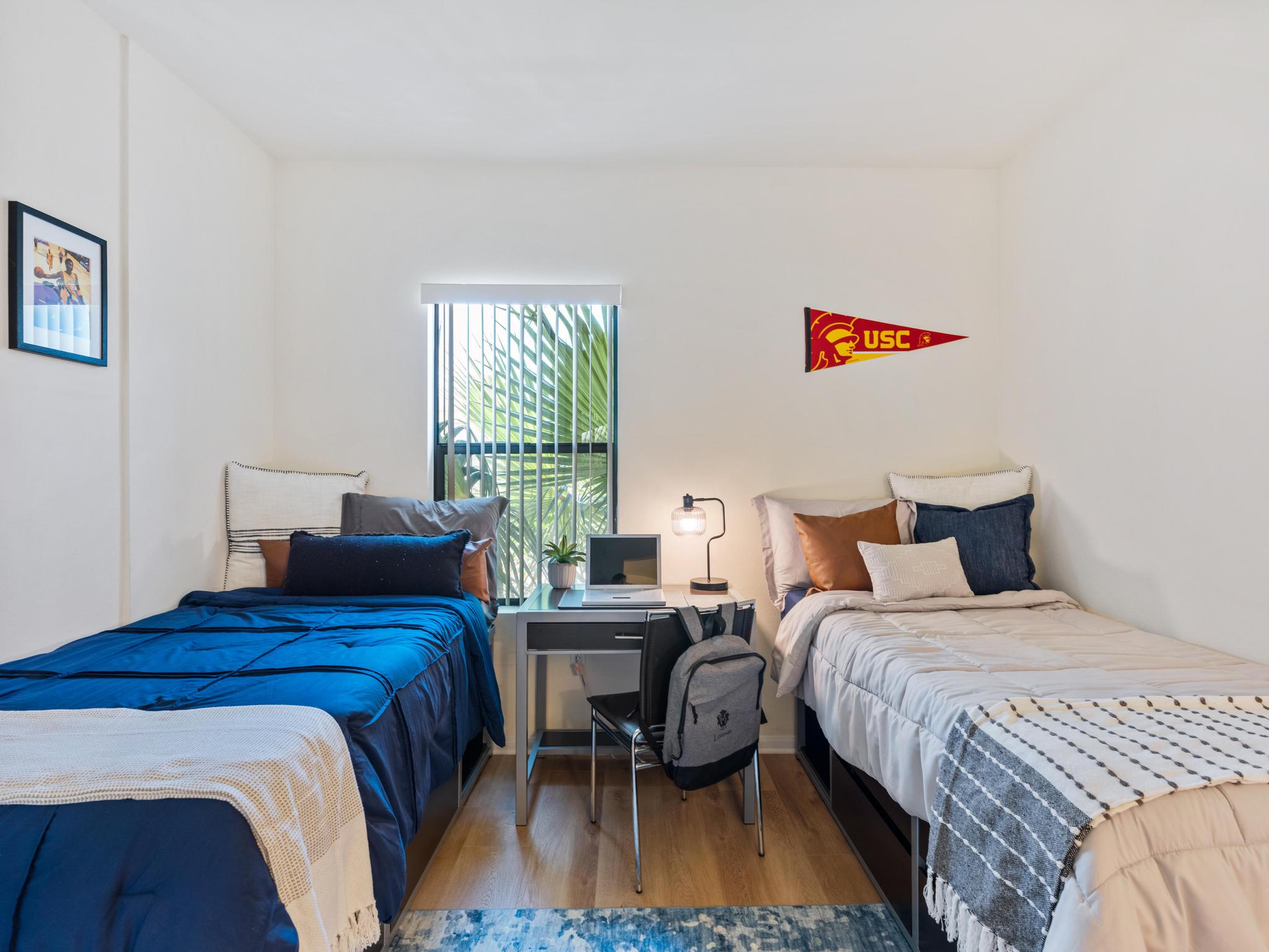 A bright and cozy bedroom featuring two twin beds with blue and neutral bedding. A small desk with a laptop and lamp is positioned between the beds. A USC pennant hangs on the wall, and a window with palm trees visible provides natural light. A backpack rests on the floor, adding to the room's student-friendly atmosphere.