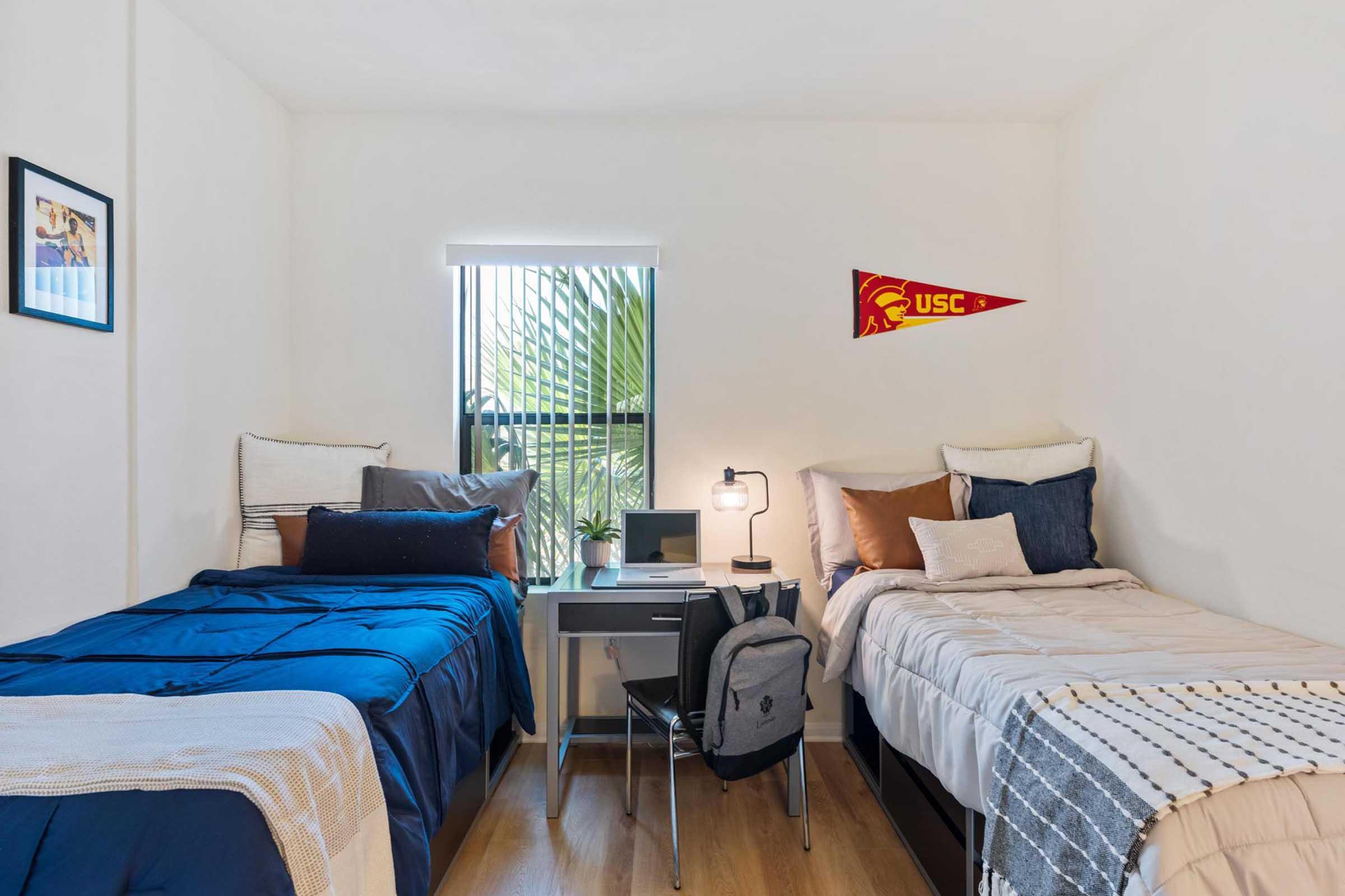 A bright, modern bedroom featuring two twin beds with blue and neutral bedding, a small desk with a lamp, and a backpack on the floor. A window allows natural light, and a USC pennant is displayed on the wall, adding a touch of school spirit. Green plants are visible outside the window.
