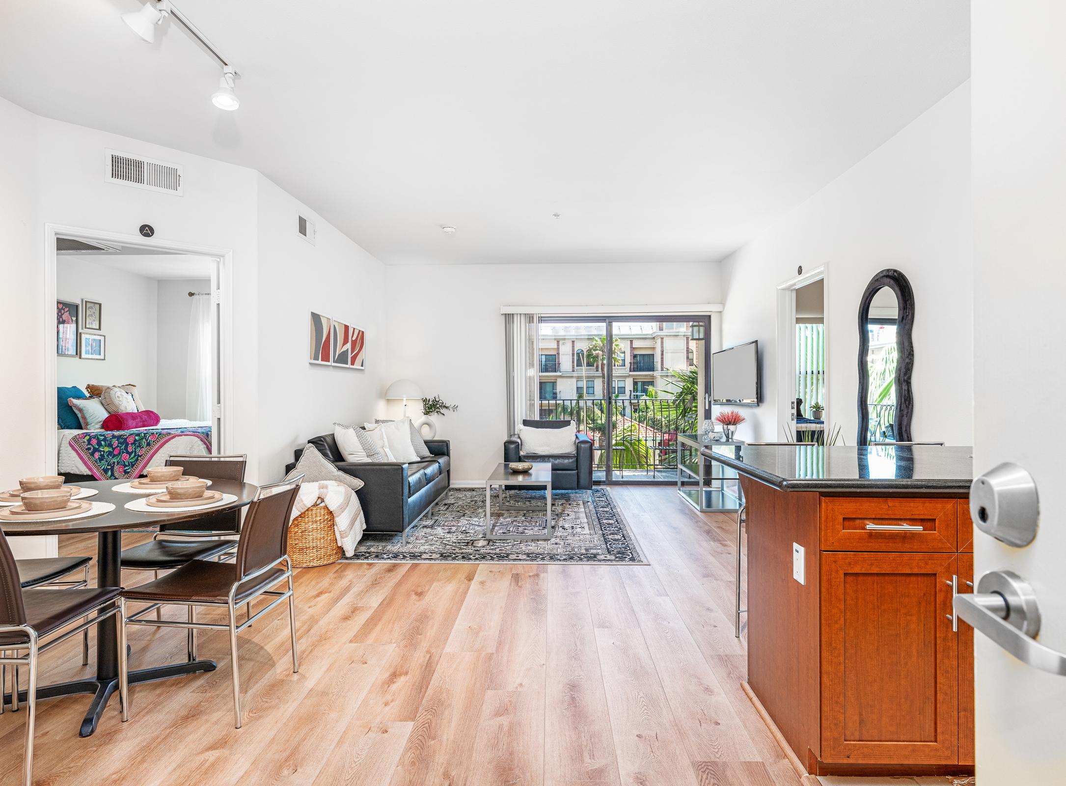 A bright and modern living space featuring a black leather sofa, a coffee table, and a dining table with chairs. The room has wood flooring and large windows that open to a patio area, allowing natural light to fill the space. Decor includes artwork and plants, creating an inviting atmosphere.