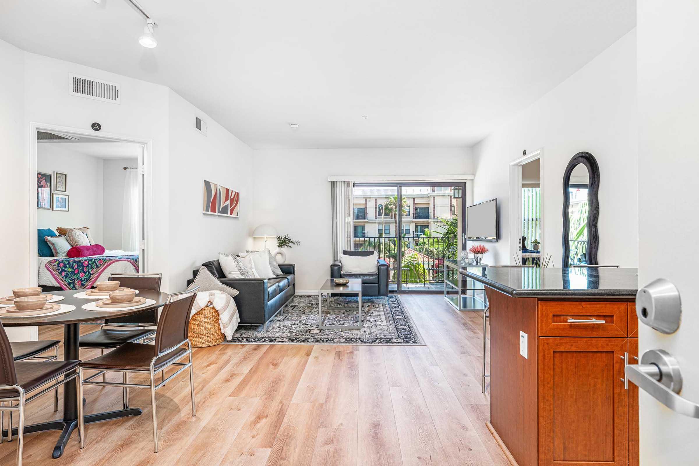 Contemporary living room with a black leather sofa, a round dining table set for four, and large windows showing a balcony with greenery. A kitchen area with wooden cabinetry is visible, and bright, modern decor adds to the inviting atmosphere.