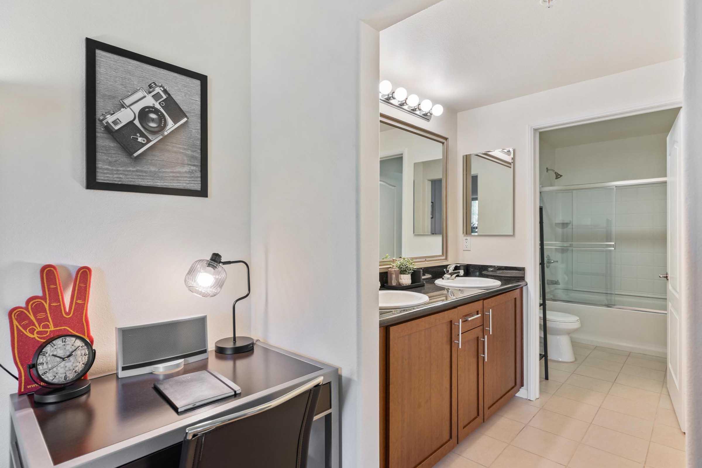 A bright and modern bathroom space with a shower and toilet visible through an open door. Adjacent to it, a small work area features a sleek desk, a stylish lamp, a clock, and a framed photo of a camera on the wall. Neutral-colored walls and light tiles contribute to the overall clean aesthetic.