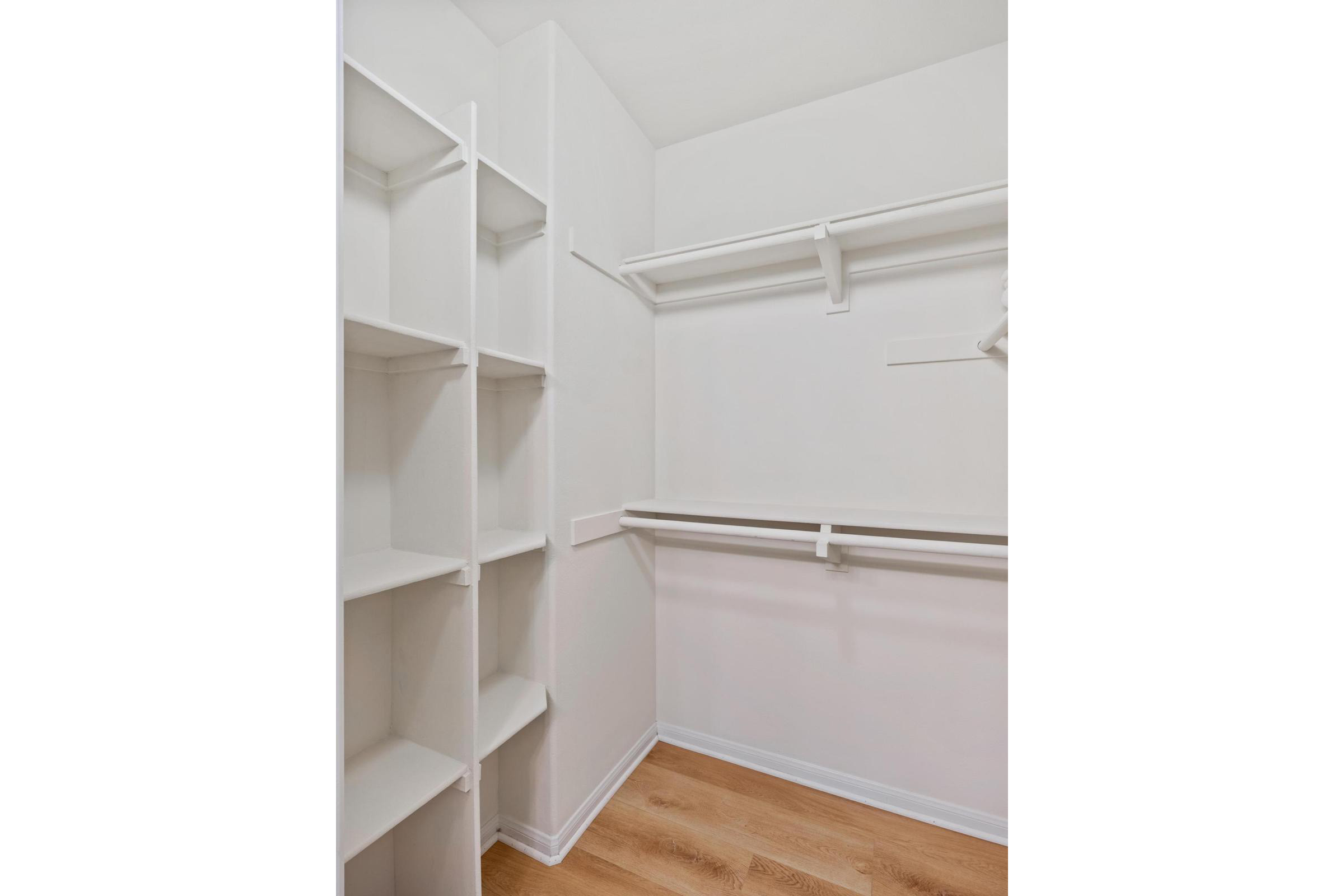 A spacious, empty walk-in closet featuring white shelving units on the left side and a hanging rod mounted on the right wall. The floor is made of light wood, and the walls are painted white, creating a bright and airy feel. The closet is uncluttered, showcasing its ample storage potential.