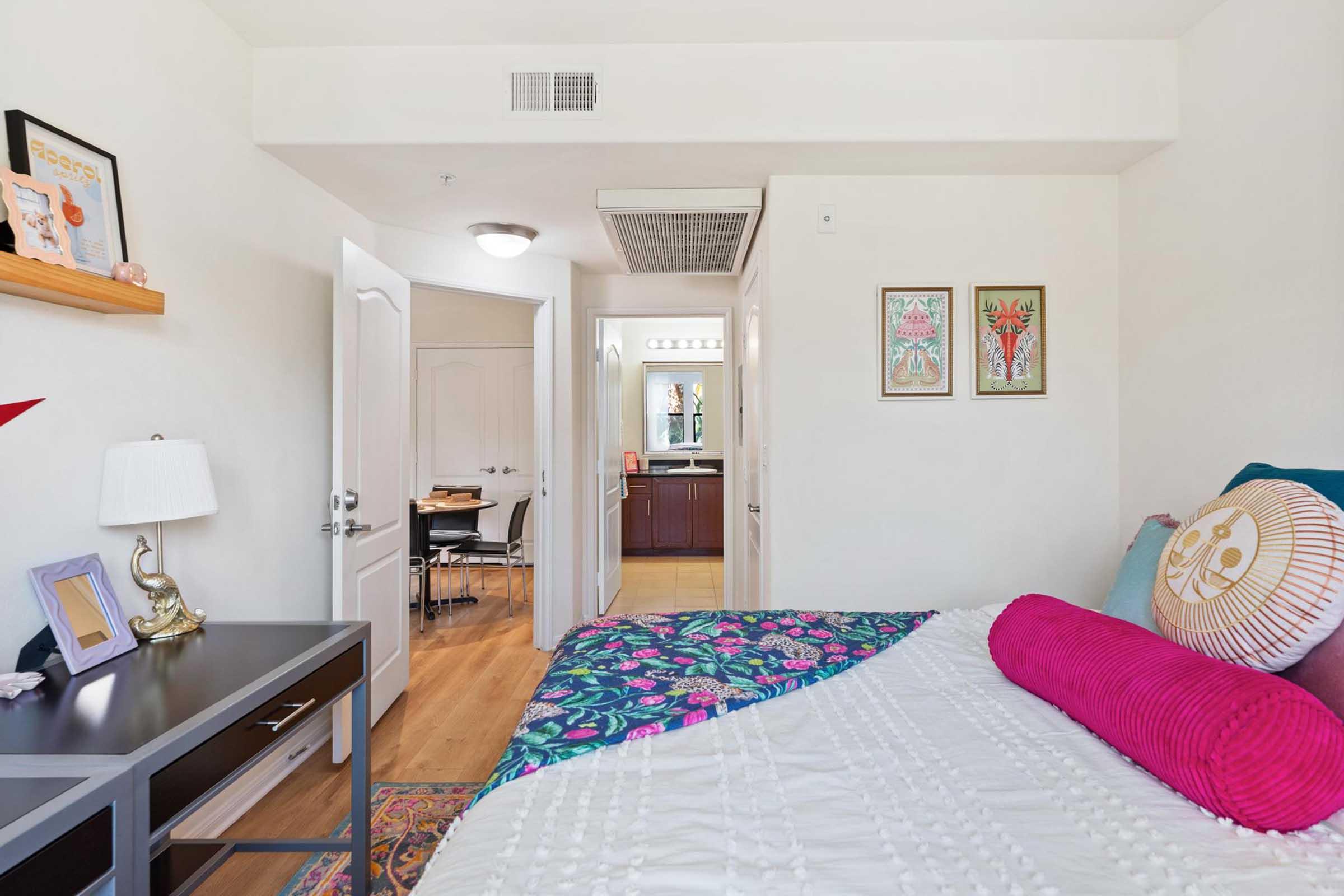 A brightly lit bedroom featuring a neatly made bed with colorful pillows, a small desk with a lamp, and decorative wall art. The room has an open doorway leading to a hallway and a kitchen area visible in the background. The decor includes floral patterns and vibrant colors, creating a cheerful atmosphere.