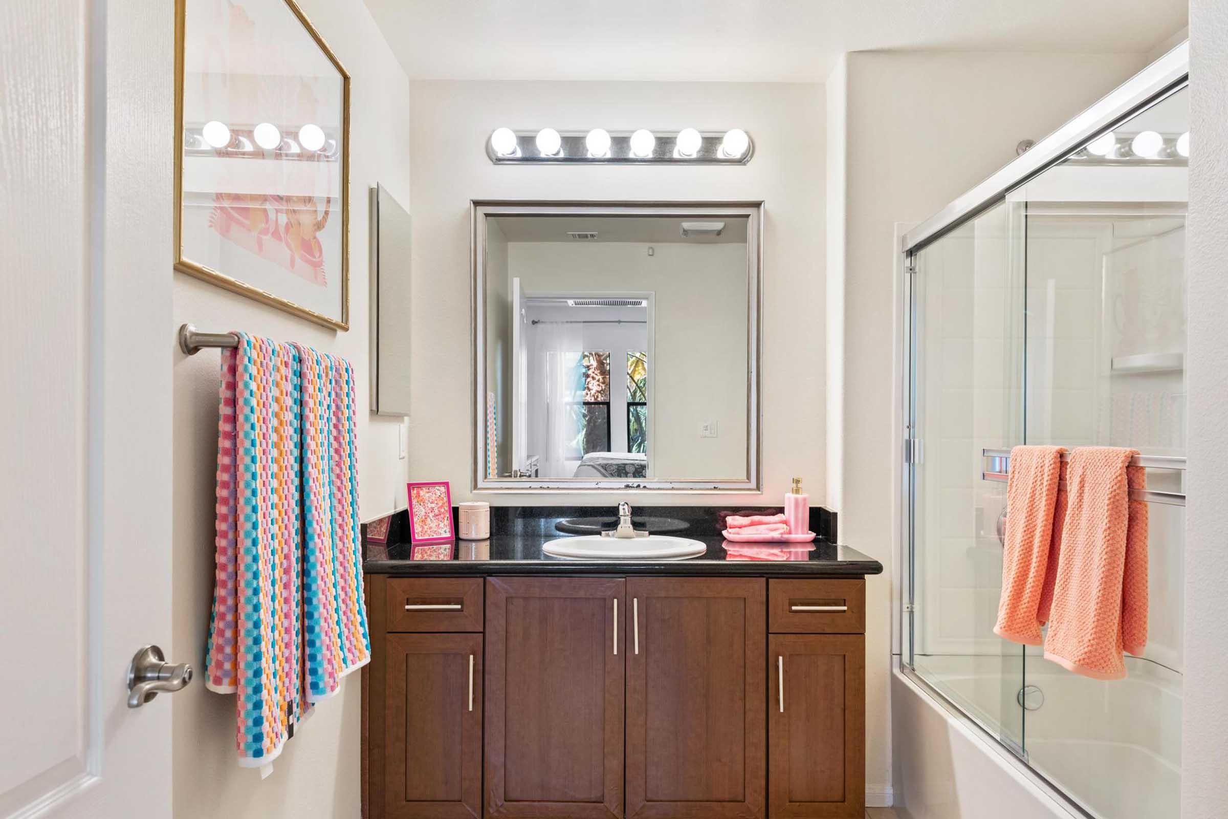 A modern bathroom featuring a dark wood vanity with a sink, a large mirror above, and a glass shower. There are colorful towels hanging on the rack and near the sink. The walls are painted in light colors, and there is a framed artwork above the vanity, adding a touch of elegance.