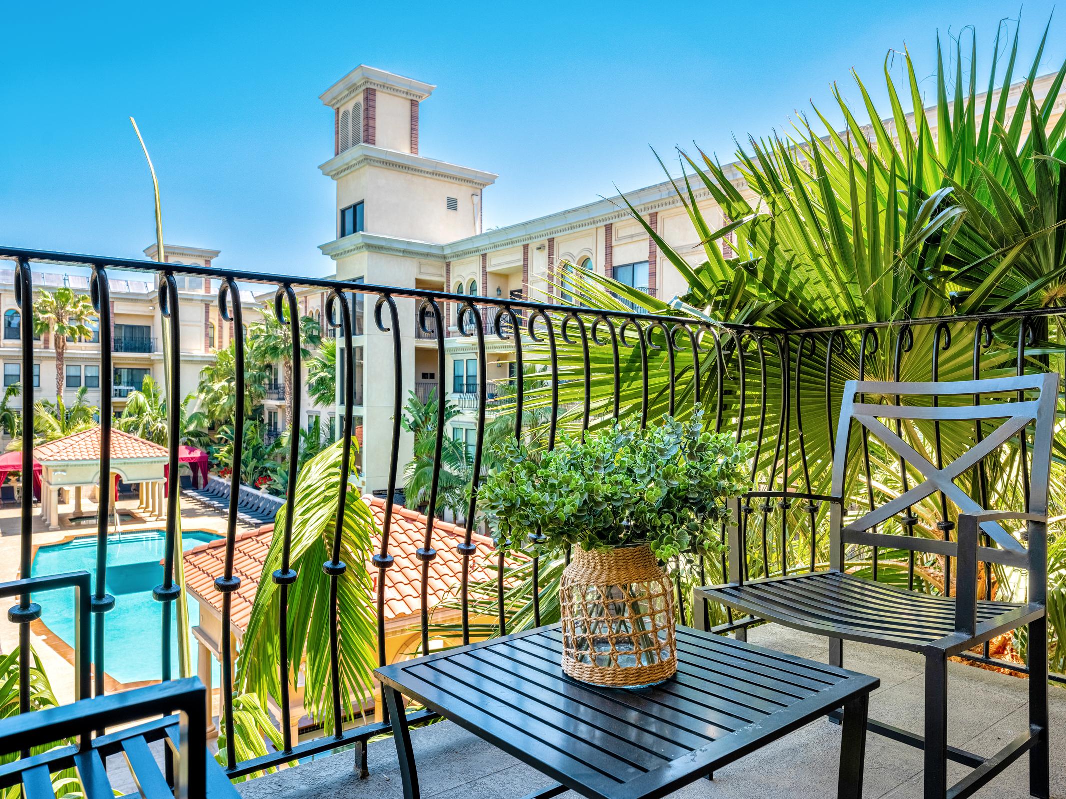 A balcony view with a black table and chair, adorned with a potted plant. In the background, a pool surrounded by lush palm trees and a building with a charming architectural design is visible, under a clear blue sky.