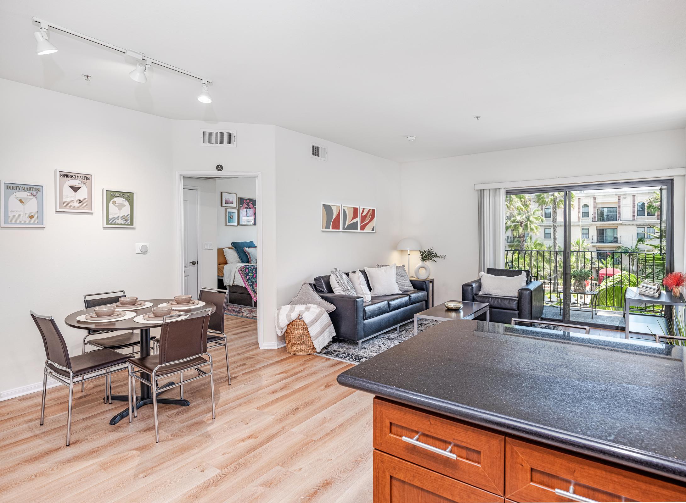 Modern apartment interior featuring a cozy living area with a sofa, chairs, and a coffee table. A dining table is set for four, and there is a large window with outdoor views. Decor includes artwork on the walls and green plants. The kitchen area is visible in the foreground with wooden cabinetry.