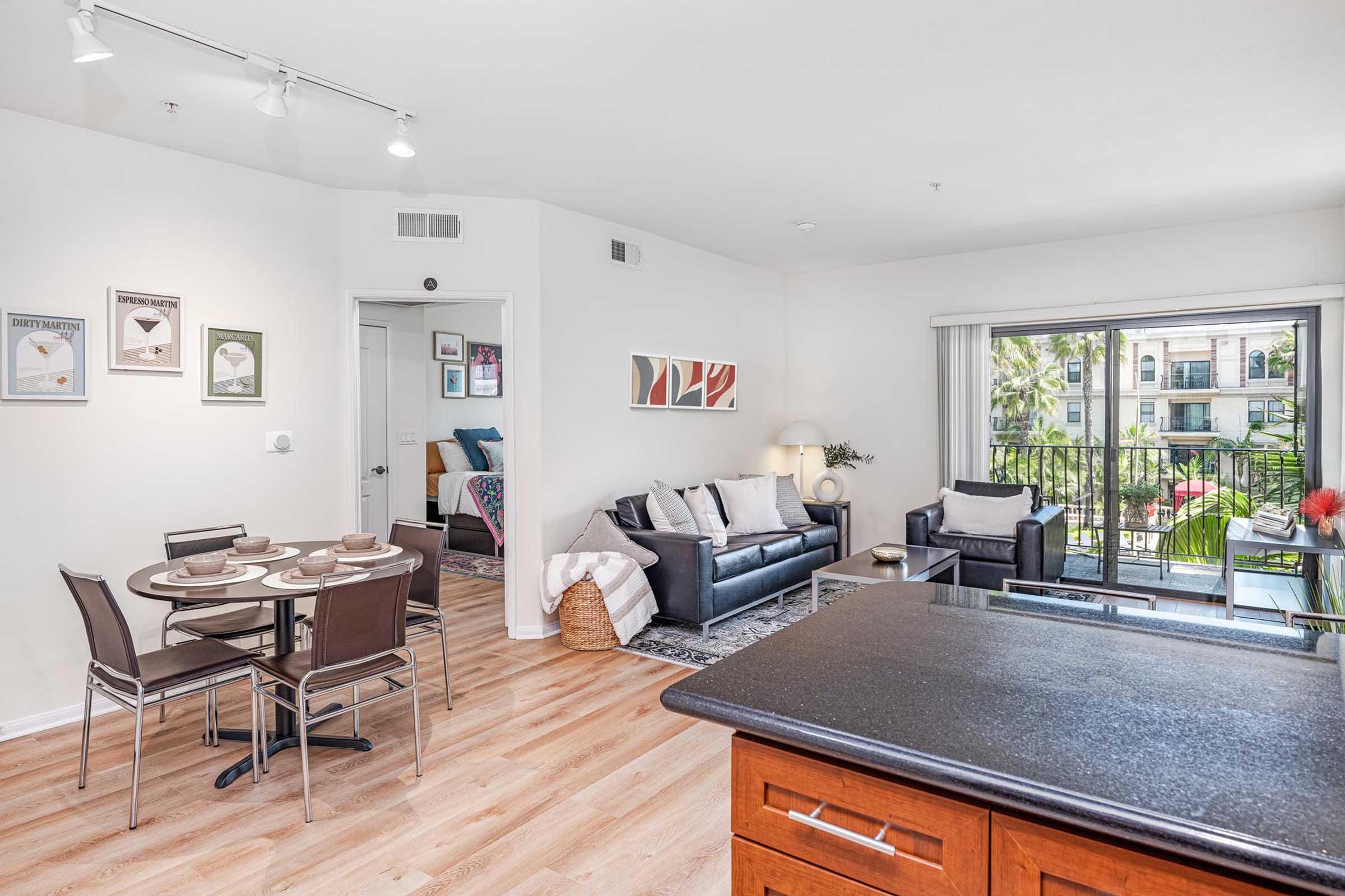 Modern apartment interior featuring a dining area with a round table set for four, a stylish living room with a sofa and armchairs, and large windows offering a view of greenery. The room has wooden flooring, decorative wall art, and contemporary furnishings, creating a bright and inviting atmosphere.