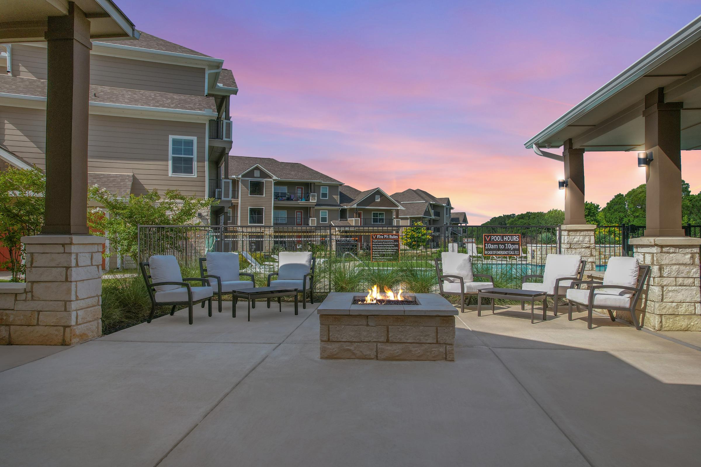 A cozy outdoor patio featuring a fire pit surrounded by comfortable chairs, with modern apartment buildings in the background. The sky is painted in soft hues of pink and purple, creating a tranquil atmosphere for relaxation and social gatherings.