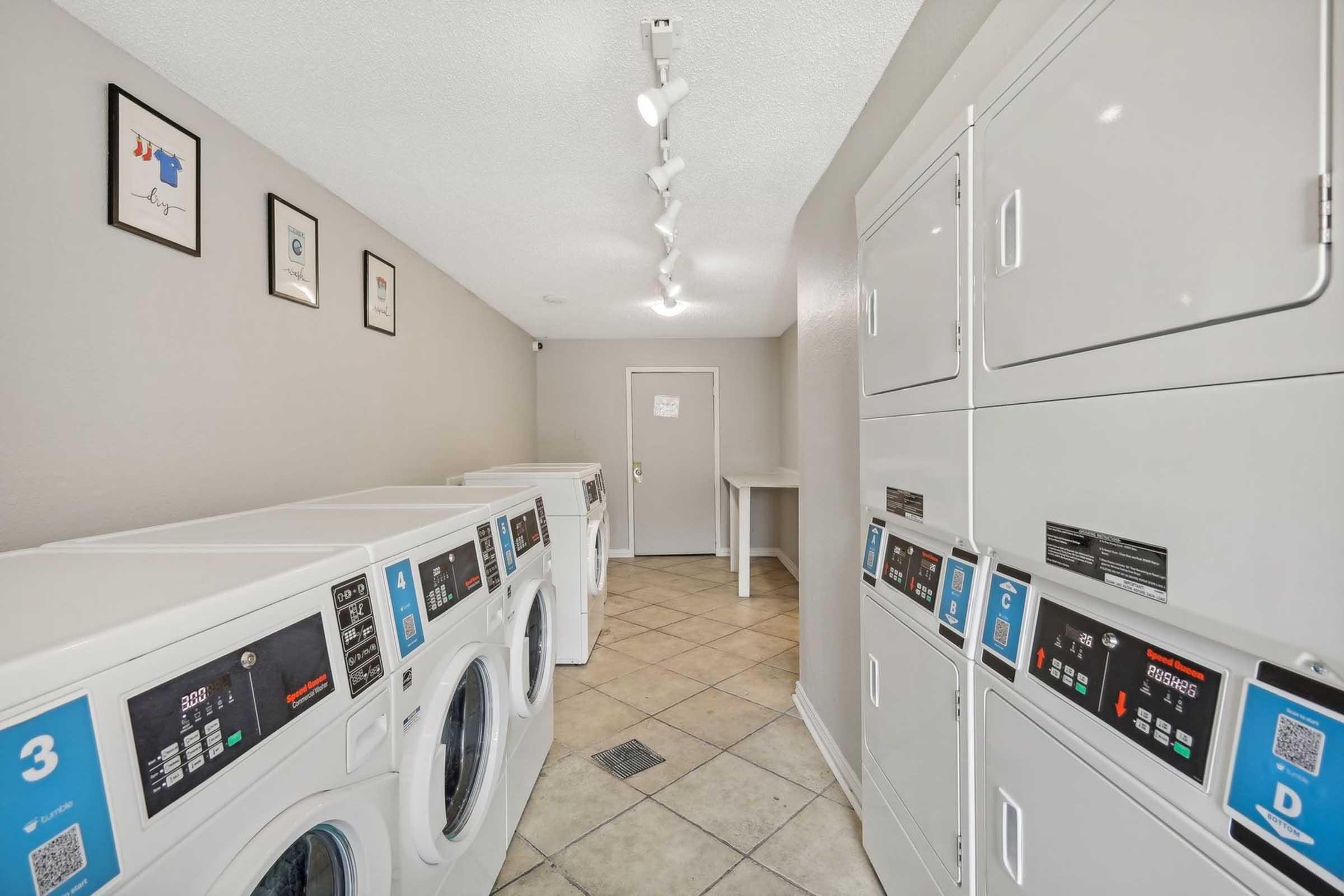 A clean and modern laundry room featuring several washing machines and dryers lined up against the wall, with a light-colored tiled floor. The walls are adorned with framed pictures, and there is a door at the end of the room. The lighting is bright, providing a welcoming atmosphere.