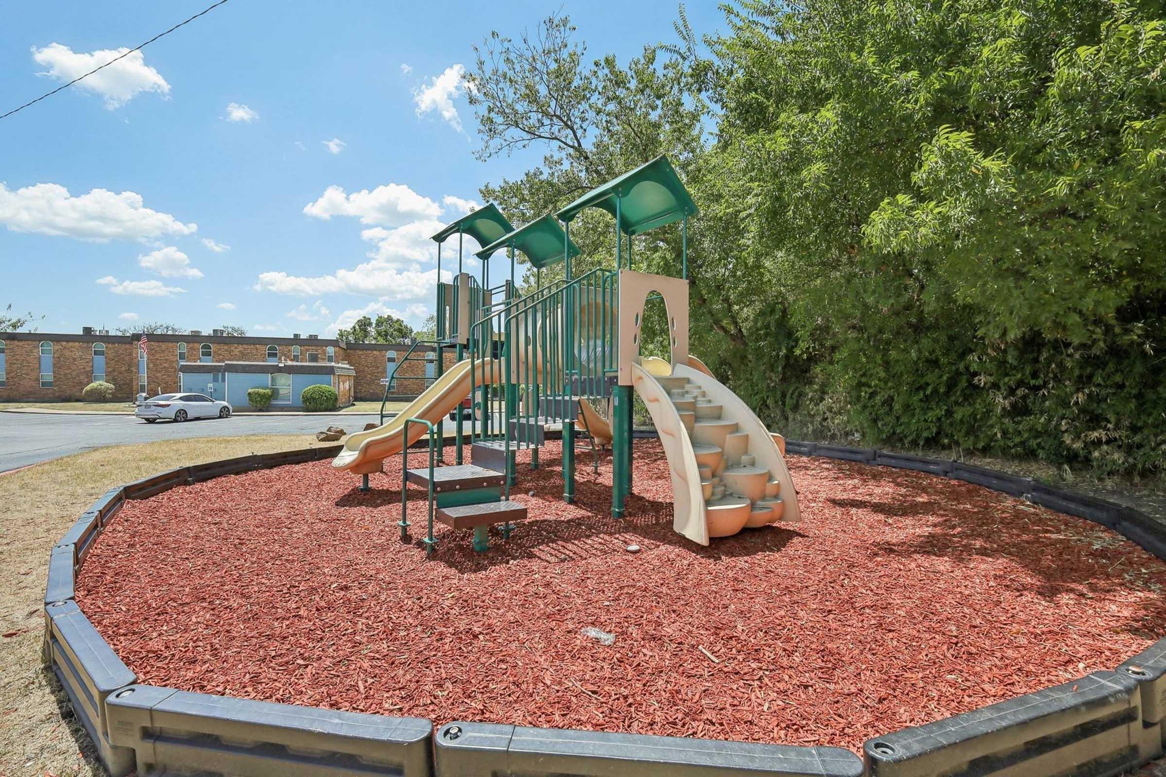 A small playground featuring a multi-level slide and climbing structure, surrounded by a circular area of red mulch. Lush green trees provide shade in the background, and a building is visible in the distance under a bright blue sky with fluffy white clouds.