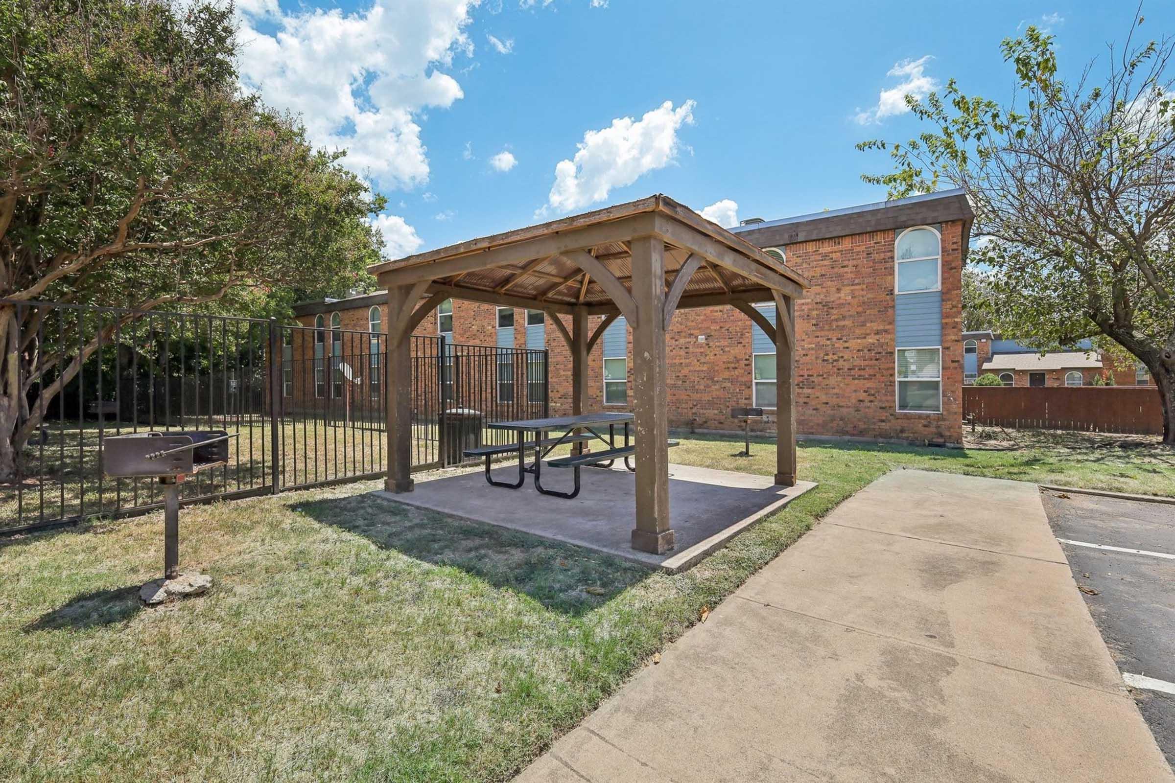 A wooden gazebo with a picnic table underneath is set on a concrete pad surrounded by a grassy area. Nearby, there's a grill and a sidewalk leading to a brick building with multiple windows. The sky is partly cloudy, creating a pleasant outdoor scene.