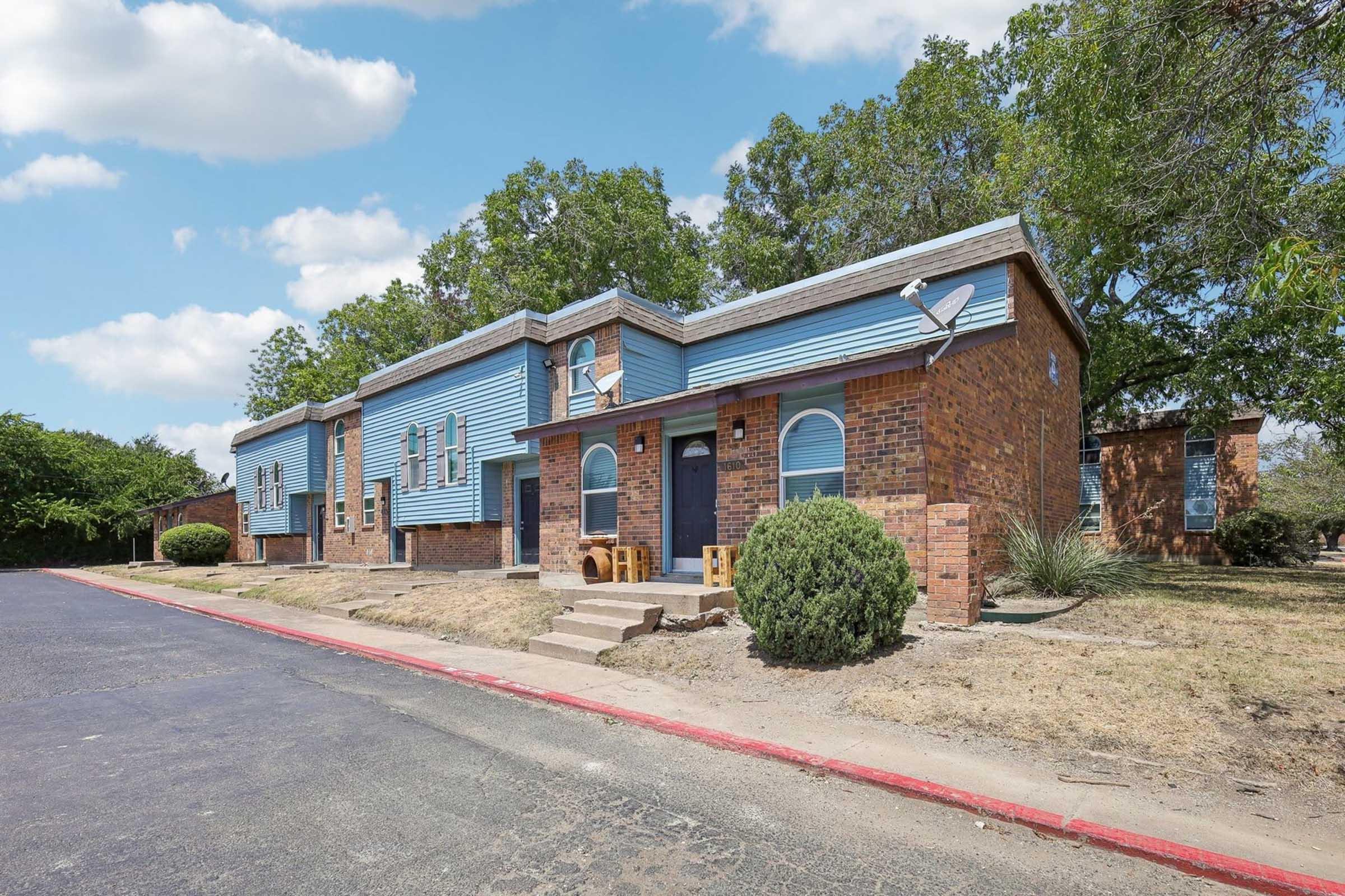 A row of brick buildings with blue siding, featuring front doors and windows. A path runs alongside the structures, lined with small shrubs. The area is partially shaded by trees under a partly cloudy sky, highlighting a suburban setting.