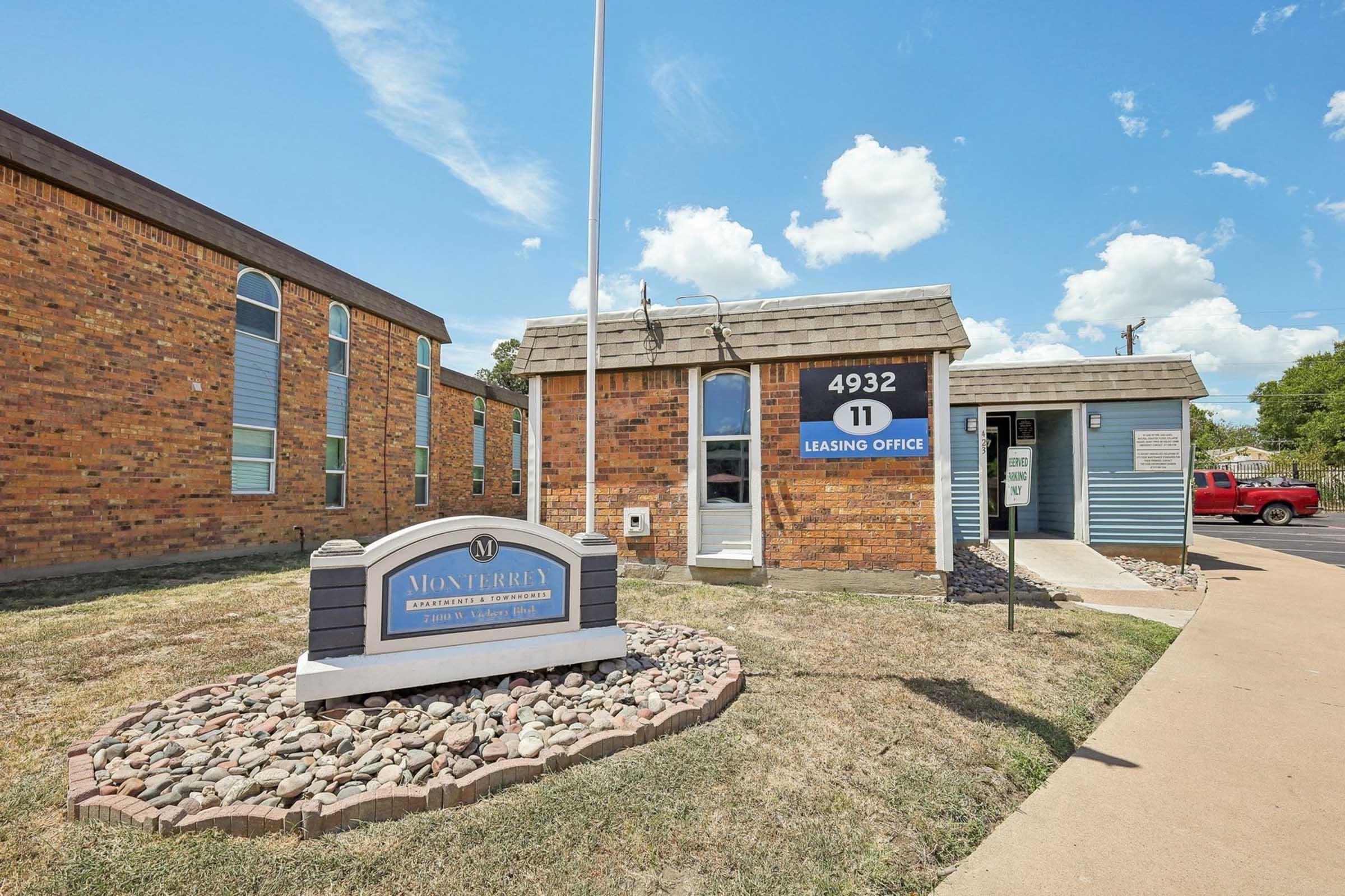 Leasing office building for Montbray Apartments, featuring a brick exterior and a blue side wall. The sign displays the address "4932" and the unit number "11." The entrance is visible, along with a well-maintained grassy area and a few decorative stones. There are fluffy clouds in a blue sky above.