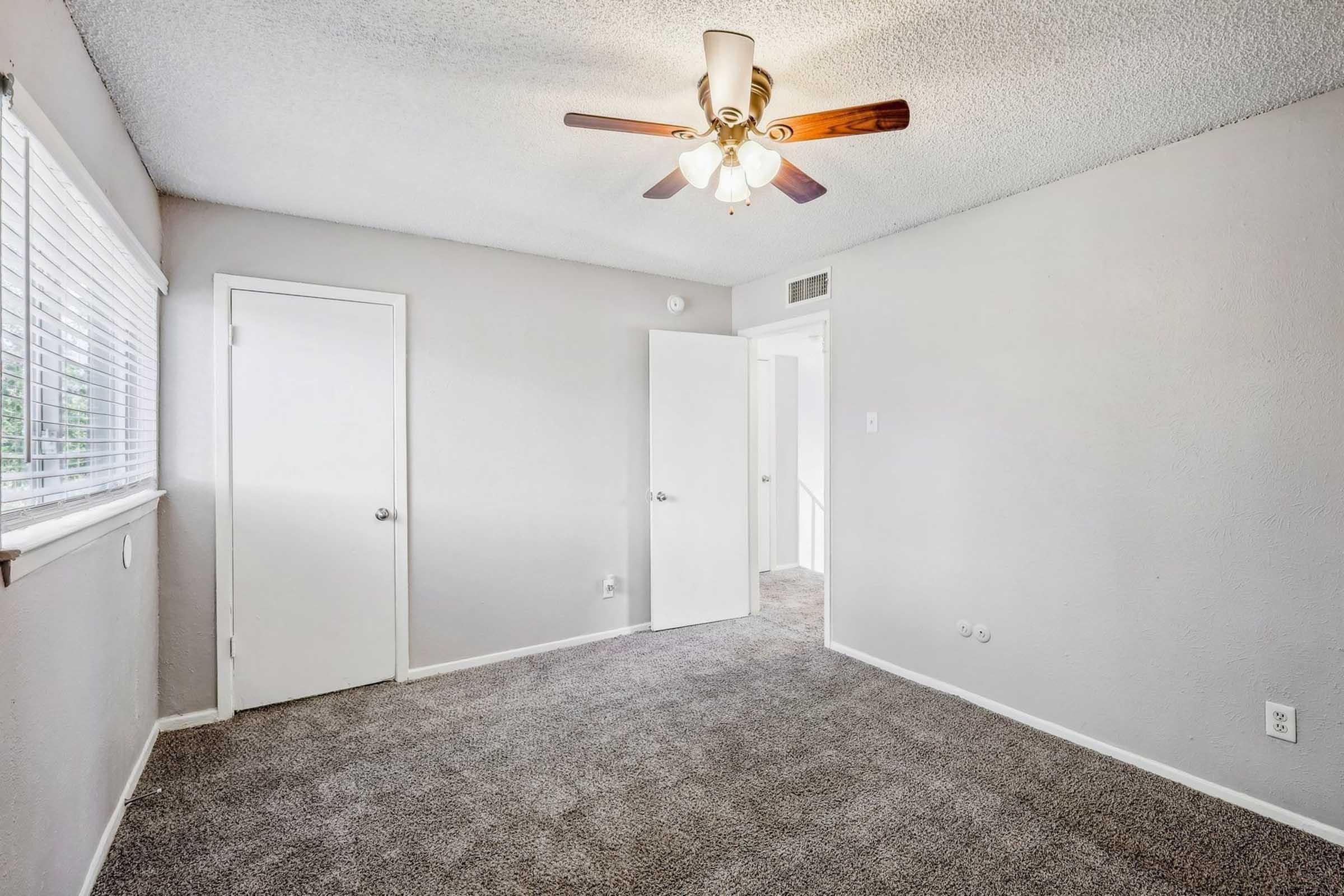 A well-lit empty room featuring a ceiling fan, light beige walls, and medium gray carpet. A window with blinds is on the left side, and two doors are visible: one closed and one partially open, leading to another area. The space appears clean and ready for furnishings.