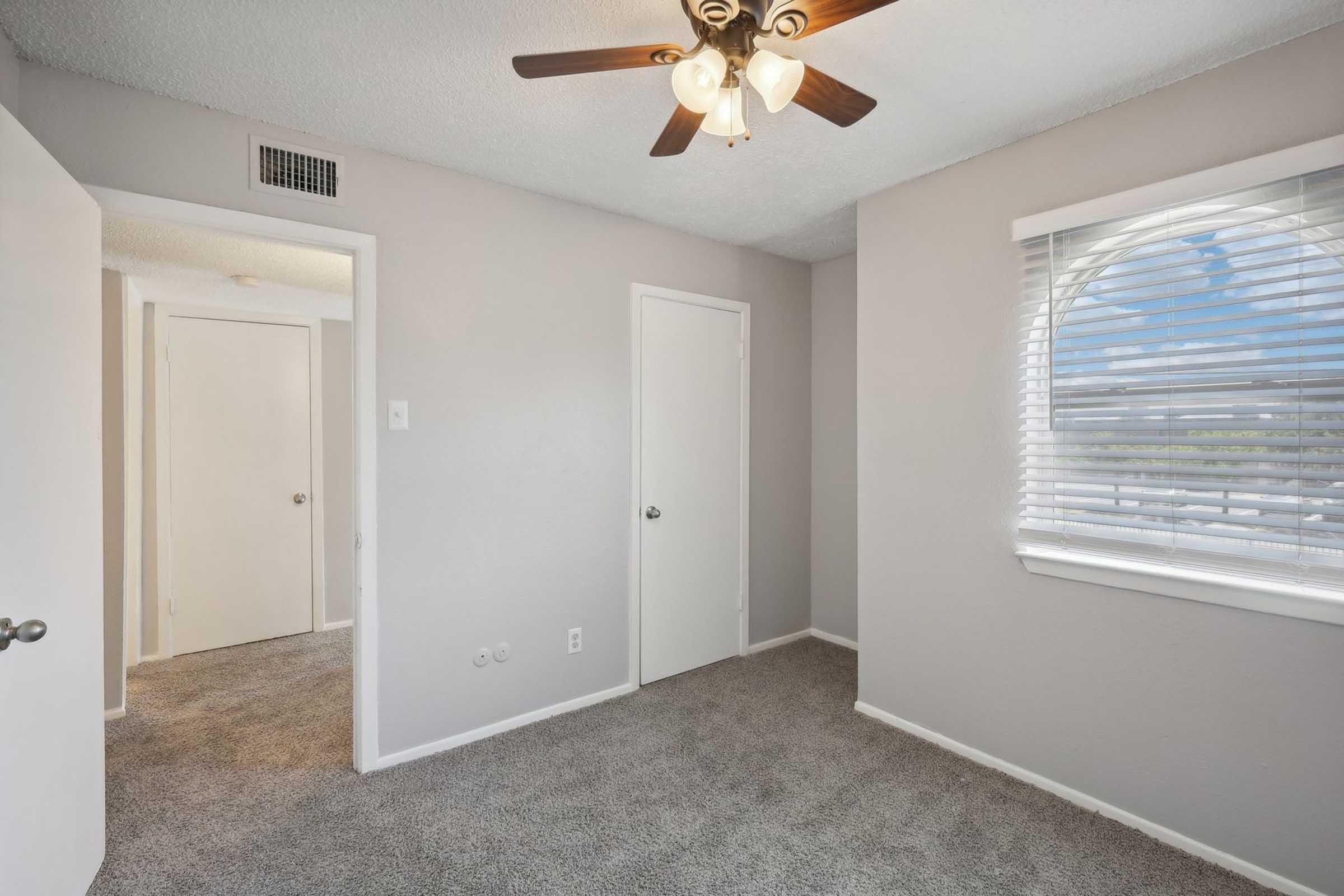 A well-lit bedroom featuring beige walls and carpet. It includes a ceiling fan with light fixtures, a large circular window with blinds, and two doors leading to closets or other rooms. The space is clean and modern, suitable for various interior design styles.
