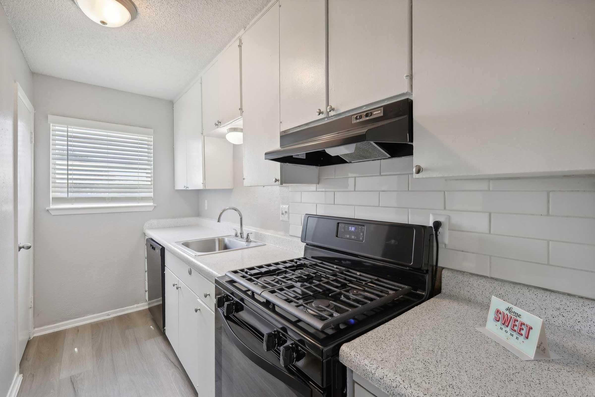 Modern kitchen featuring a black gas stove, white cabinets, and a sink. The countertops are light gray with a speckled pattern. A window with blinds allows natural light in, and a small sign on the countertop reads "SWEET." The overall color scheme is light and contemporary.