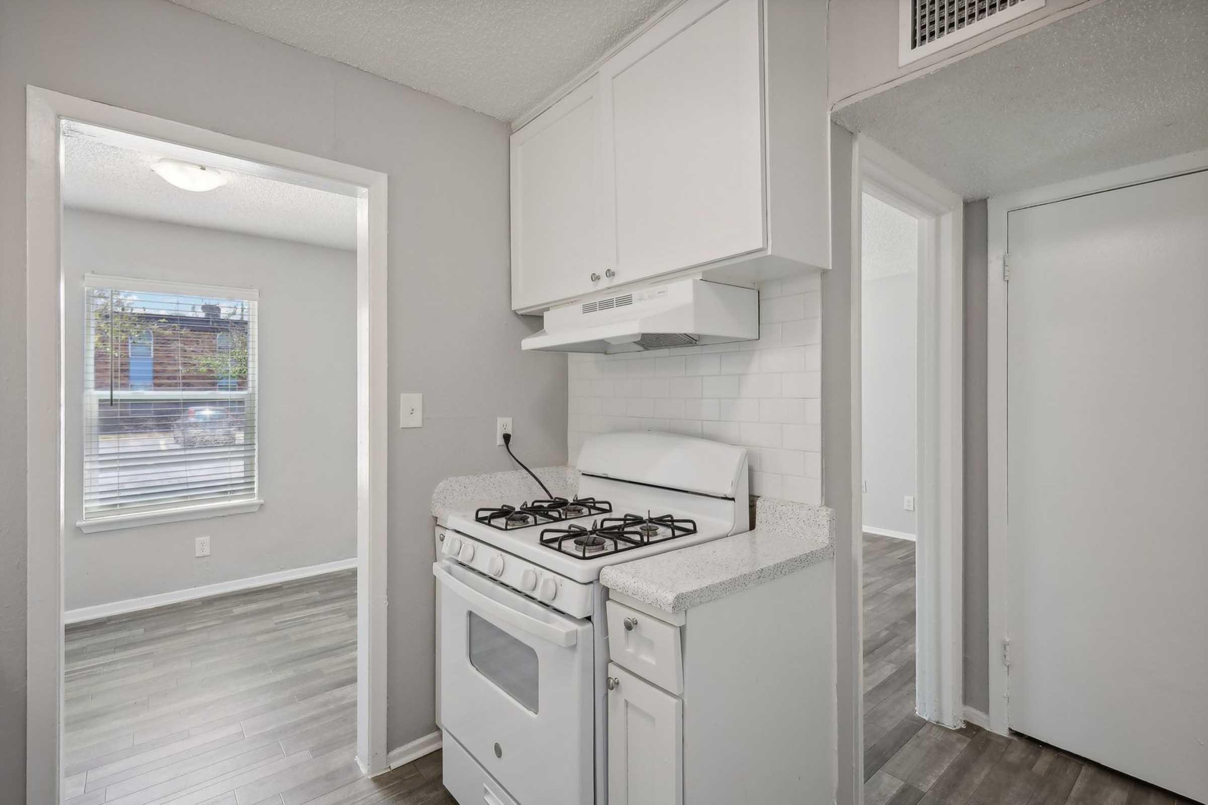 A clean, modern kitchen featuring a white gas stove, white cabinetry, and a light-colored countertop. Adjacent is a doorway leading to another room, with a window providing natural light. The floor is covered with grey vinyl planks, and the walls are painted in a soft grey tone.