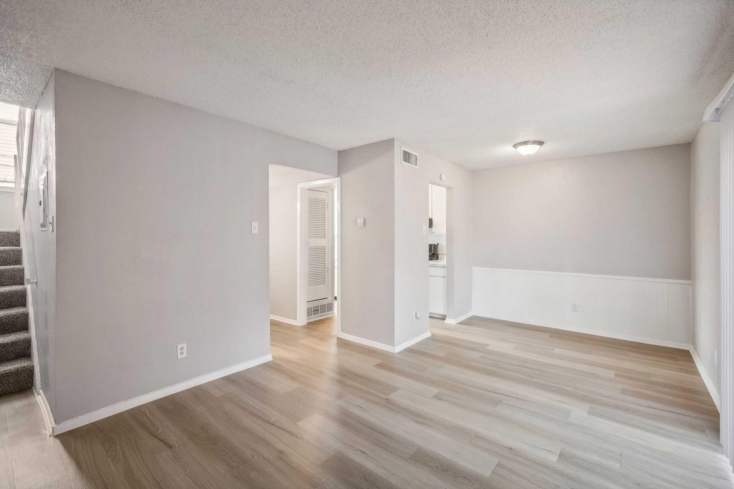Spacious, light-filled room featuring light grey walls, warm wood flooring, and a ceiling light fixture. A doorway leads to a small kitchen area, while a staircase is visible in the background. Perfect for a cozy living space or dining area.