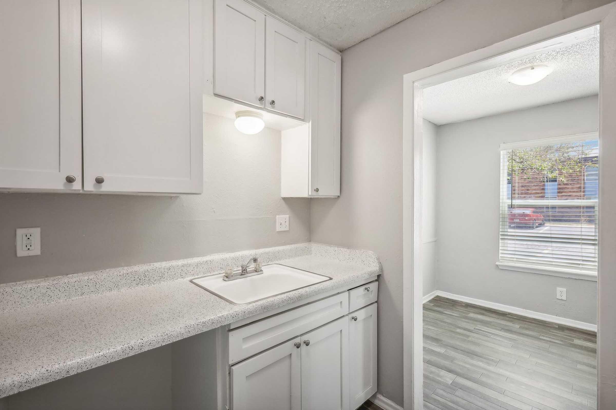 A small kitchen area featuring white cabinets, a sink, and a light-colored countertop. There is a doorway leading to a bright room with a window and gray flooring. Natural light is coming through the window, creating a cozy atmosphere.
