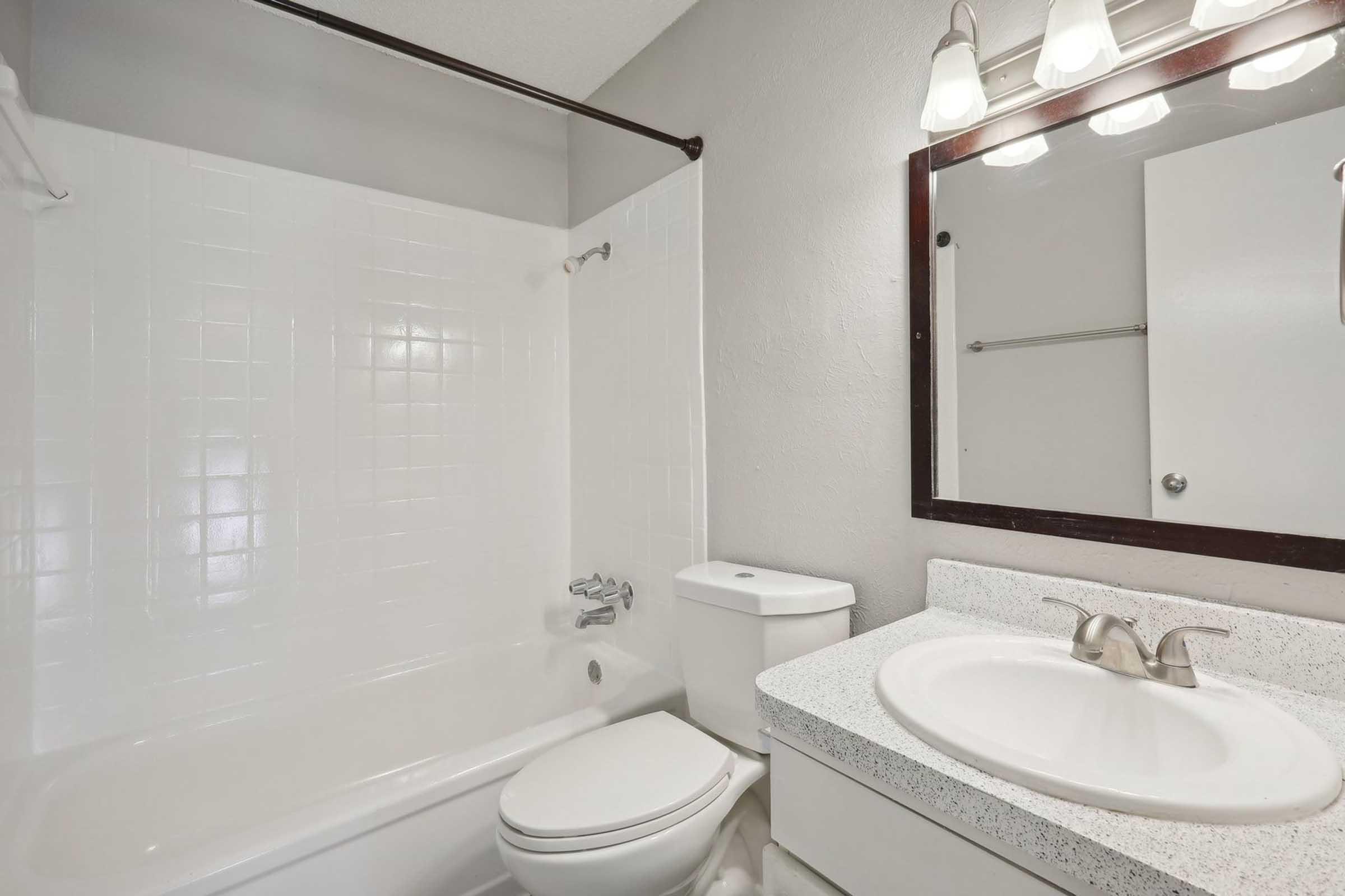 A clean, modern bathroom featuring a white bathtub with a shower, a toilet, and a sink with a countertop. The walls are painted in a light gray color, and there is a mirror above the sink with three light fixtures above it. The ambiance is bright and inviting.