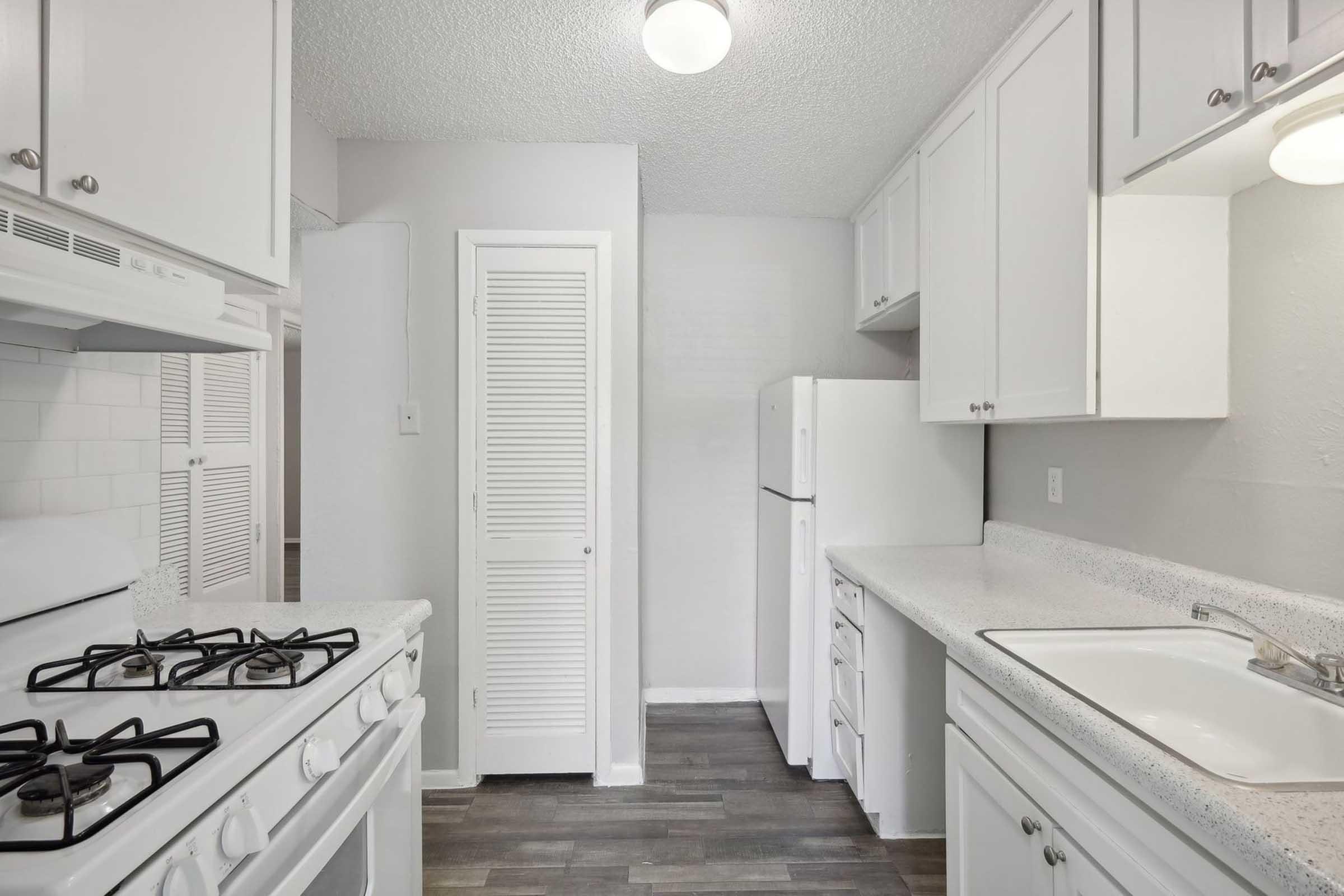 A modern, well-lit kitchen featuring white cabinets, a gas stove, a refrigerator, and a spacious countertop. There is a sink with a faucet, and the flooring is dark laminate. The walls are painted a light color, creating an open and inviting atmosphere.