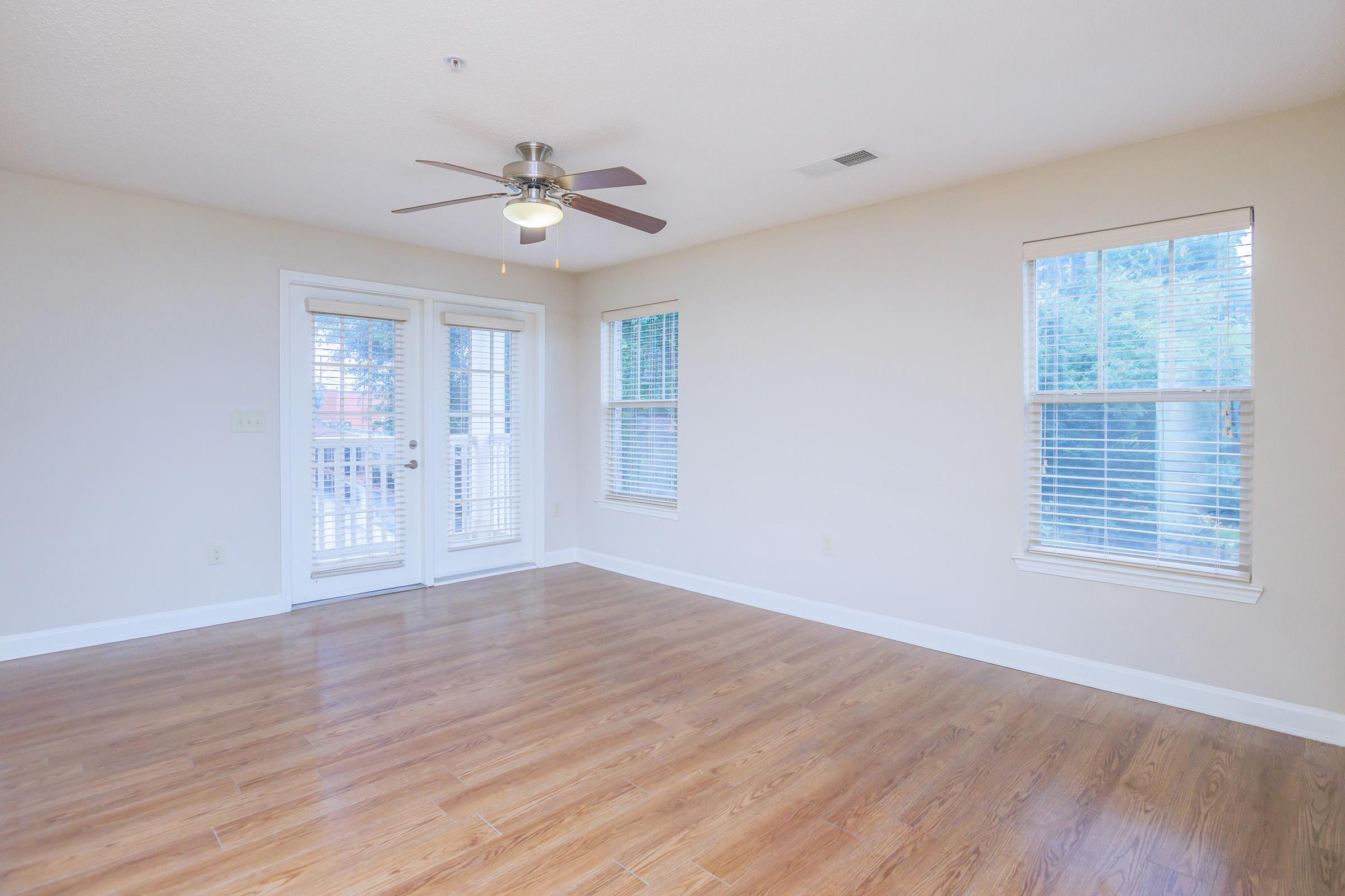 A spacious, empty room with light brown wooden flooring and a ceiling fan. Two large windows with blinds allow natural light to enter, and there are glass doors leading to an outdoor area. The walls are painted a light color, creating a bright and inviting atmosphere.