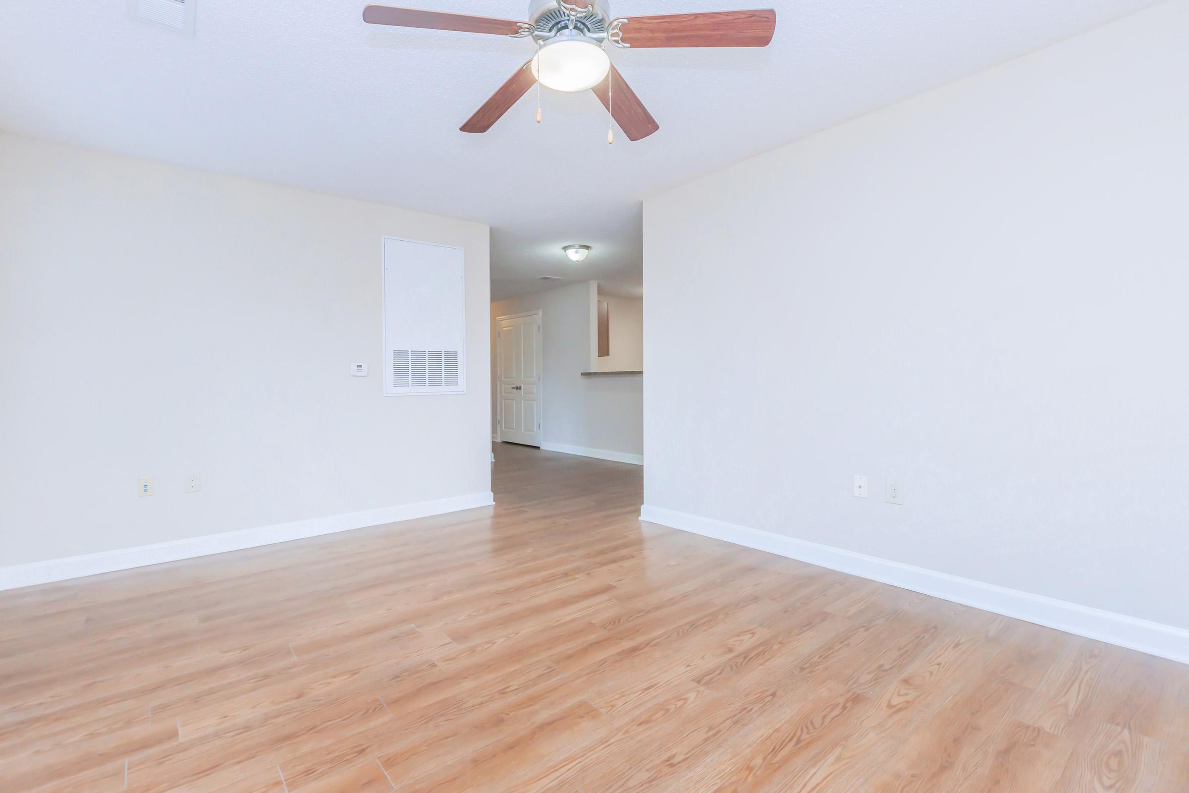 Bright, empty room with light-colored walls and polished hardwood flooring. A ceiling fan with wooden blades hangs from the ceiling, and an air vent is visible on the wall. In the background, a doorway leads to a kitchen area, creating an open layout feel.
