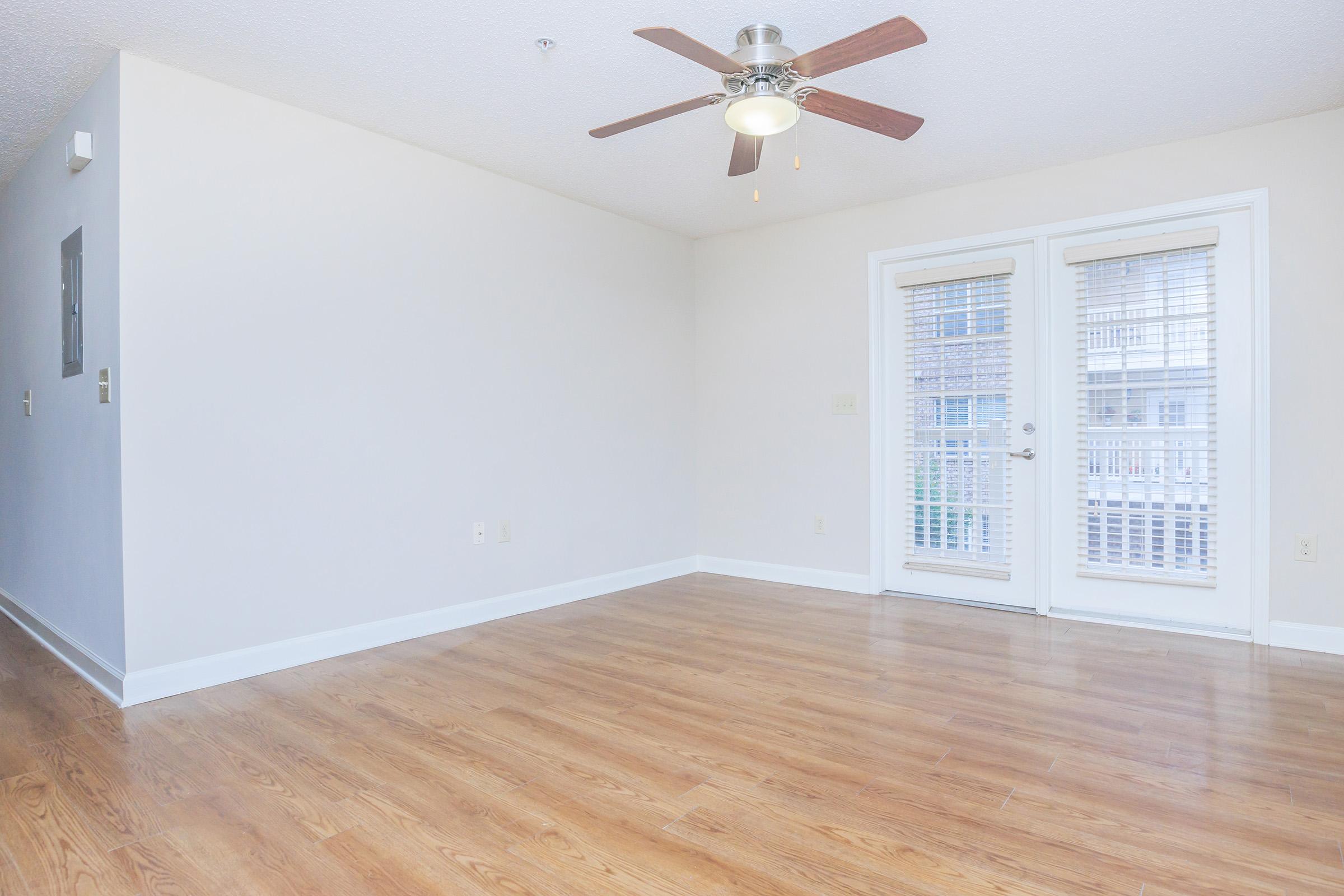 A spacious, empty room with light wooden flooring and a ceiling fan. Two glass doors lead to a balcony, allowing natural light to enter. The walls are painted in a soft, neutral color, and there are no furnishings or decorations, emphasizing the room's openness.