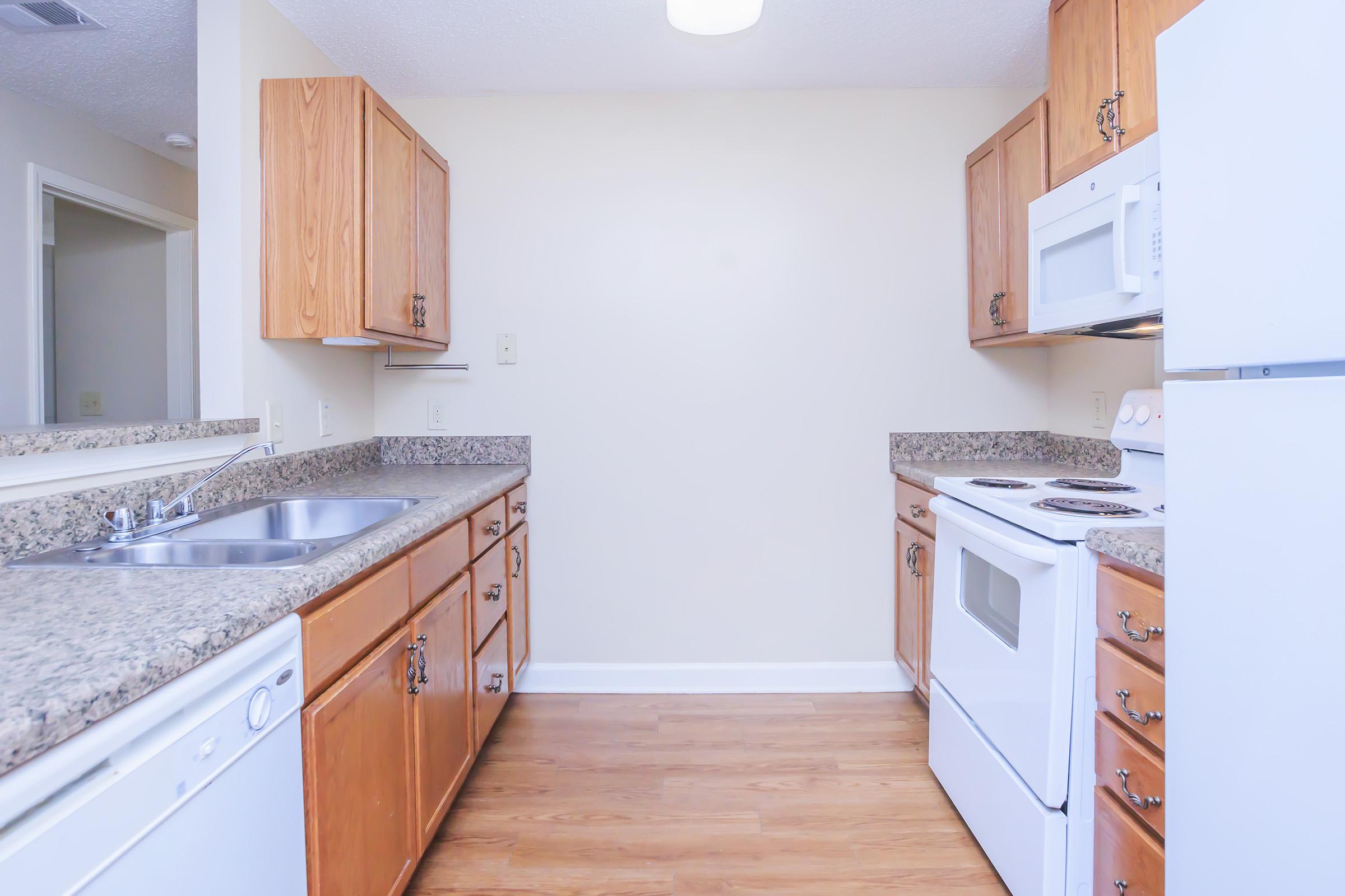 A modern kitchen featuring light wood cabinets, granite countertops, and stainless steel appliances. The space includes a double sink, dishwasher, electric stove, and microwave, with a neutral-colored wall and wooden flooring enhancing the brightness of the room. Ideal for cooking and dining.