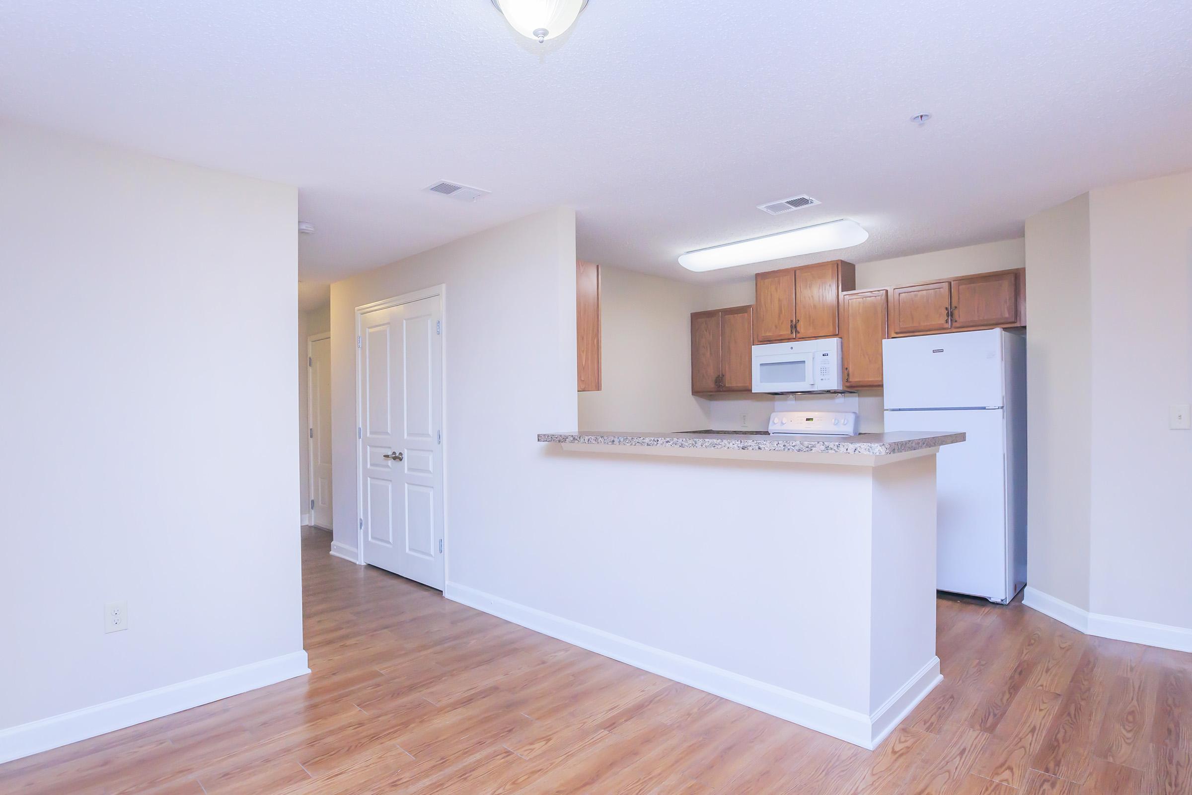A view of a spacious, well-lit kitchen and living area in an apartment. The kitchen features wooden cabinets, a white microwave, and a refrigerator, with a granite bar counter separating it from the living space. Light hardwood flooring extends throughout the area, with neutral-colored walls and bright overhead lighting.