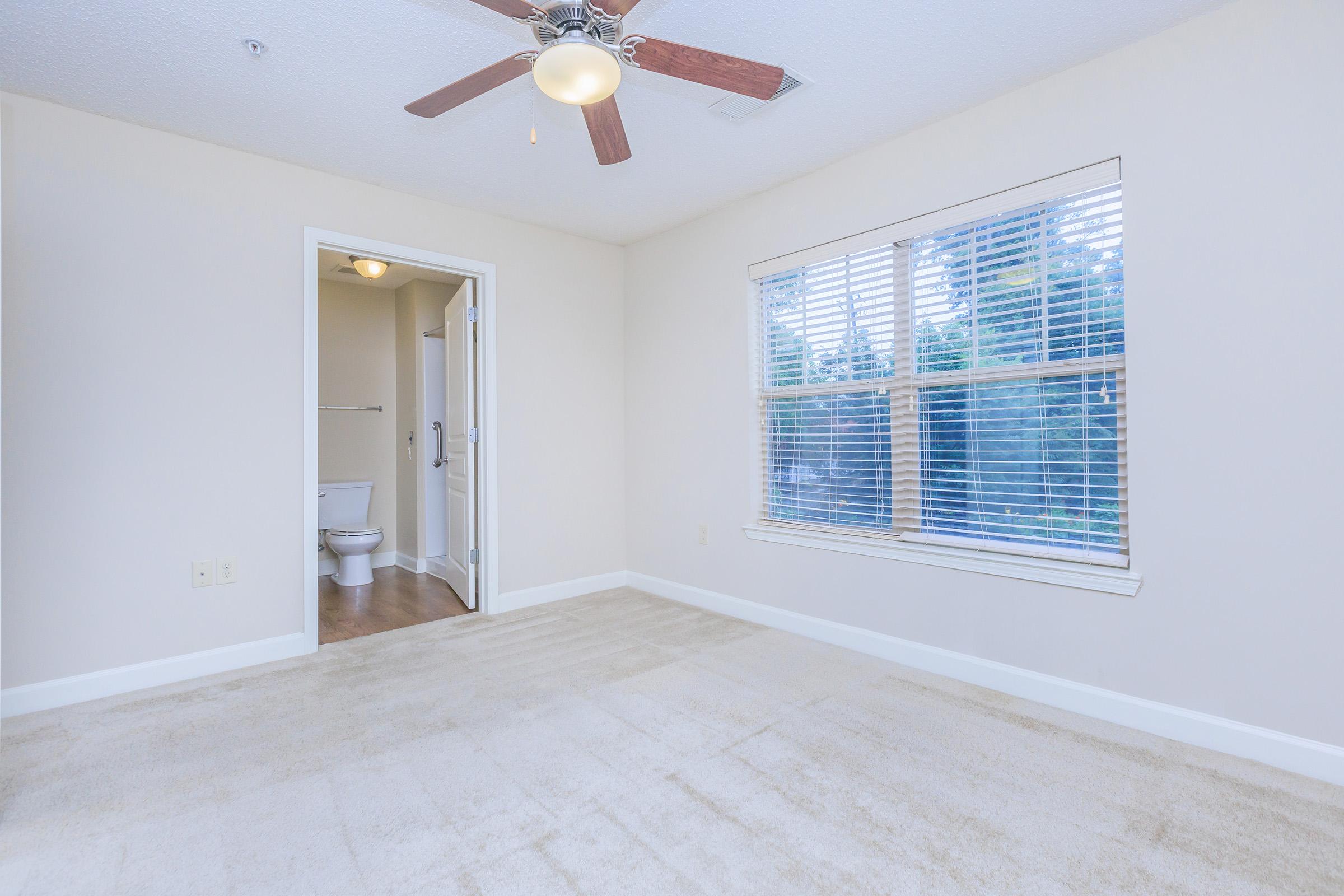 A bright, empty room with light-colored carpet and walls. A ceiling fan hangs from the ceiling, and a window with blinds allows natural light in. There is an open doorway leading to a bathroom with a toilet visible. The overall atmosphere is spacious and clean.