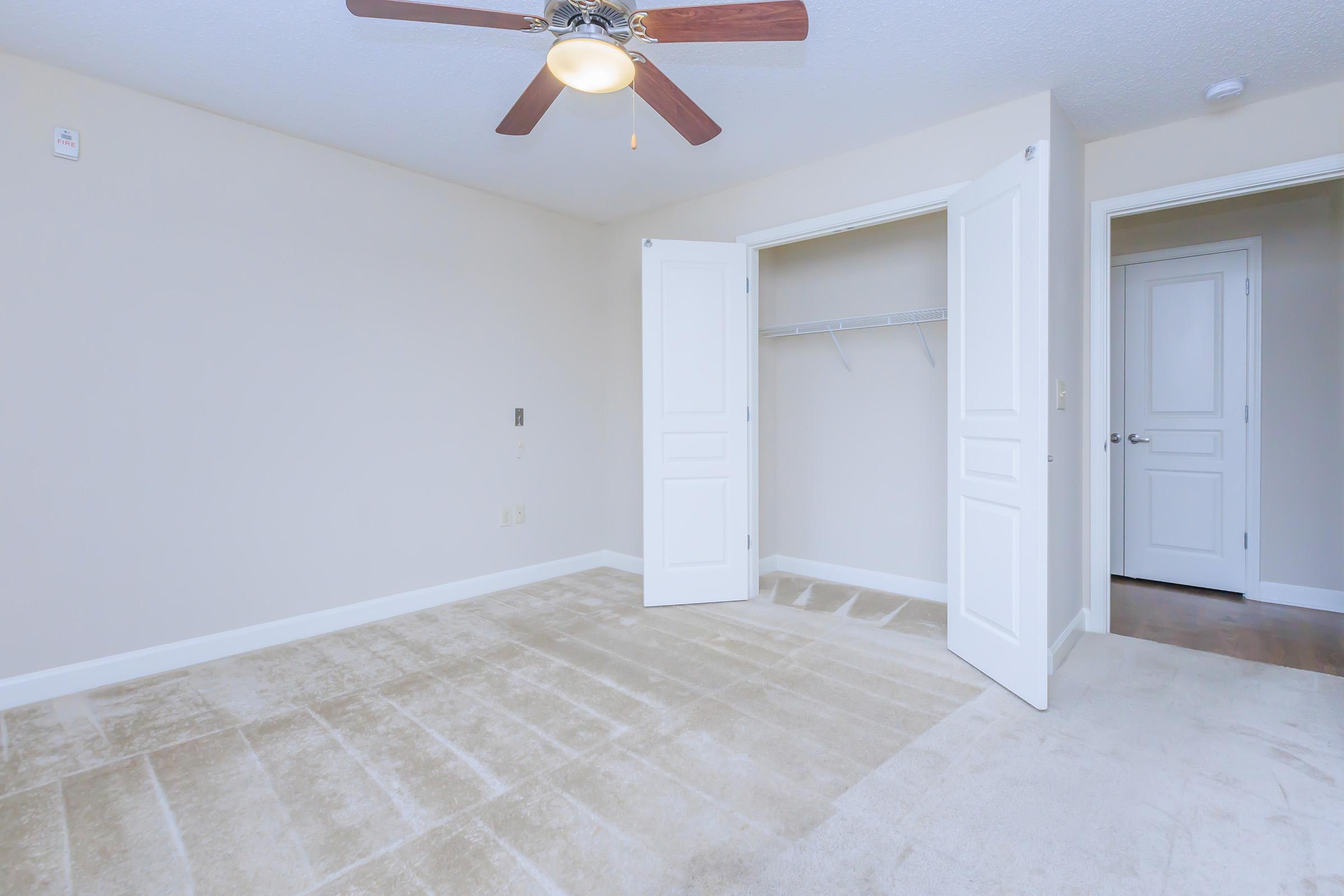 Empty room with light beige walls, a ceiling fan, and a plush beige carpet. The room features a double closet with white doors open, revealing a shelf inside. A door on the right leads to another area, completing the simple and spacious layout. Natural light fills the space, creating a bright and airy atmosphere.