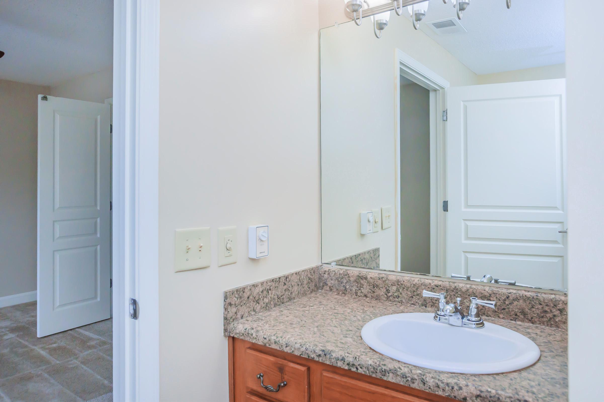 A bright bathroom with a light-colored granite countertop and a sink. The mirror reflects the room, showing a white door in the background. The walls are painted in a neutral tone, and the flooring features square tiles. The lighting is soft and natural, creating a welcoming atmosphere.