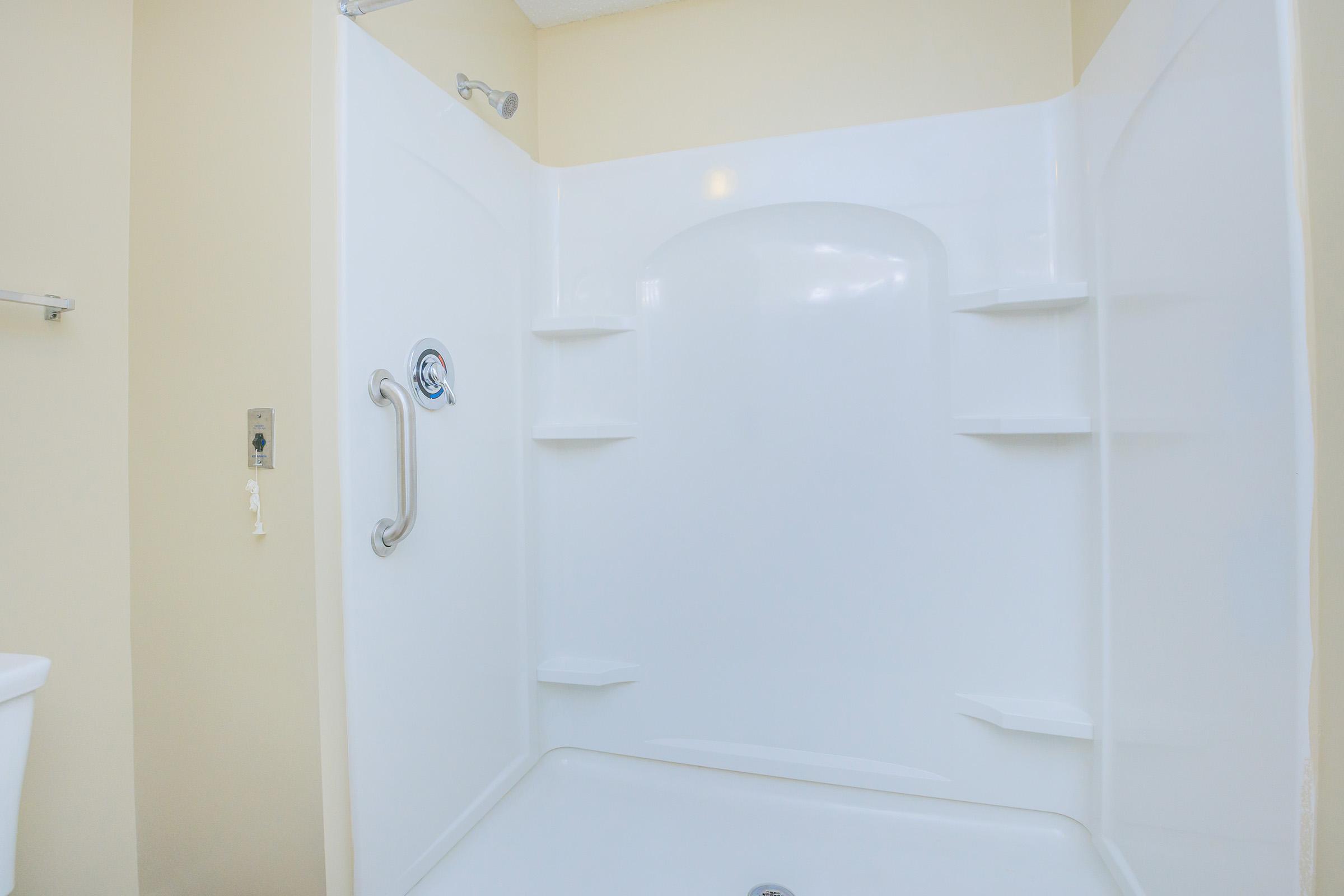 A clean, white shower stall with smooth walls and built-in shelves. The shower features a grab bar for safety and a handheld showerhead. The background is a light-colored wall, suggesting a bright and airy bathroom space.
