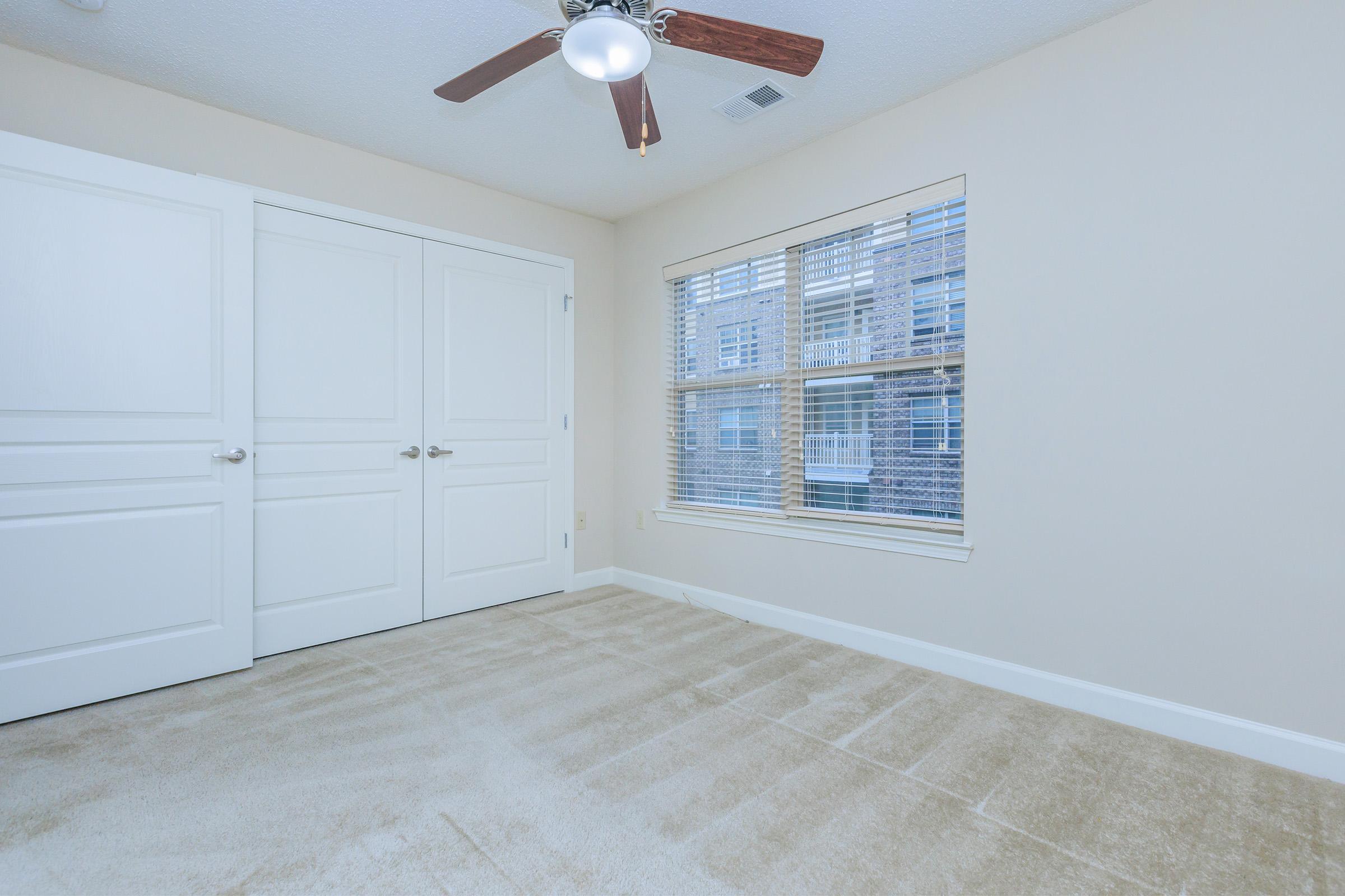 An empty, well-lit room featuring beige walls and a carpeted floor. There is a window with blinds allowing natural light to enter, and a ceiling fan with wooden blades. Two white doors are visible, suggesting storage or closet space, while the overall ambiance is clean and minimalist.