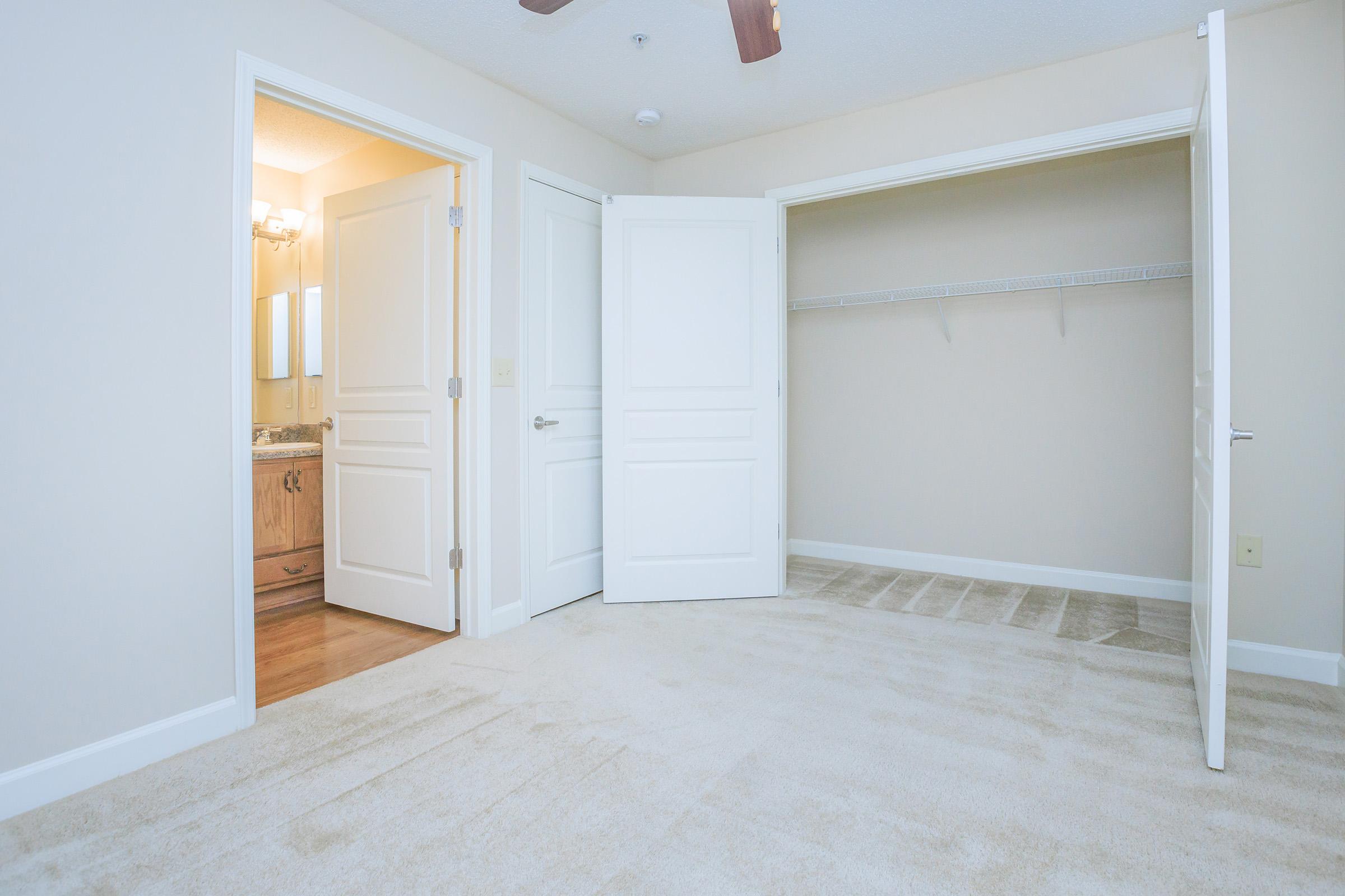 A spacious, empty room with light-colored walls and carpet. Two white doors are open, revealing an adjoining bathroom and a closet with a shelf. Soft lighting illuminates the area, creating a bright atmosphere.
