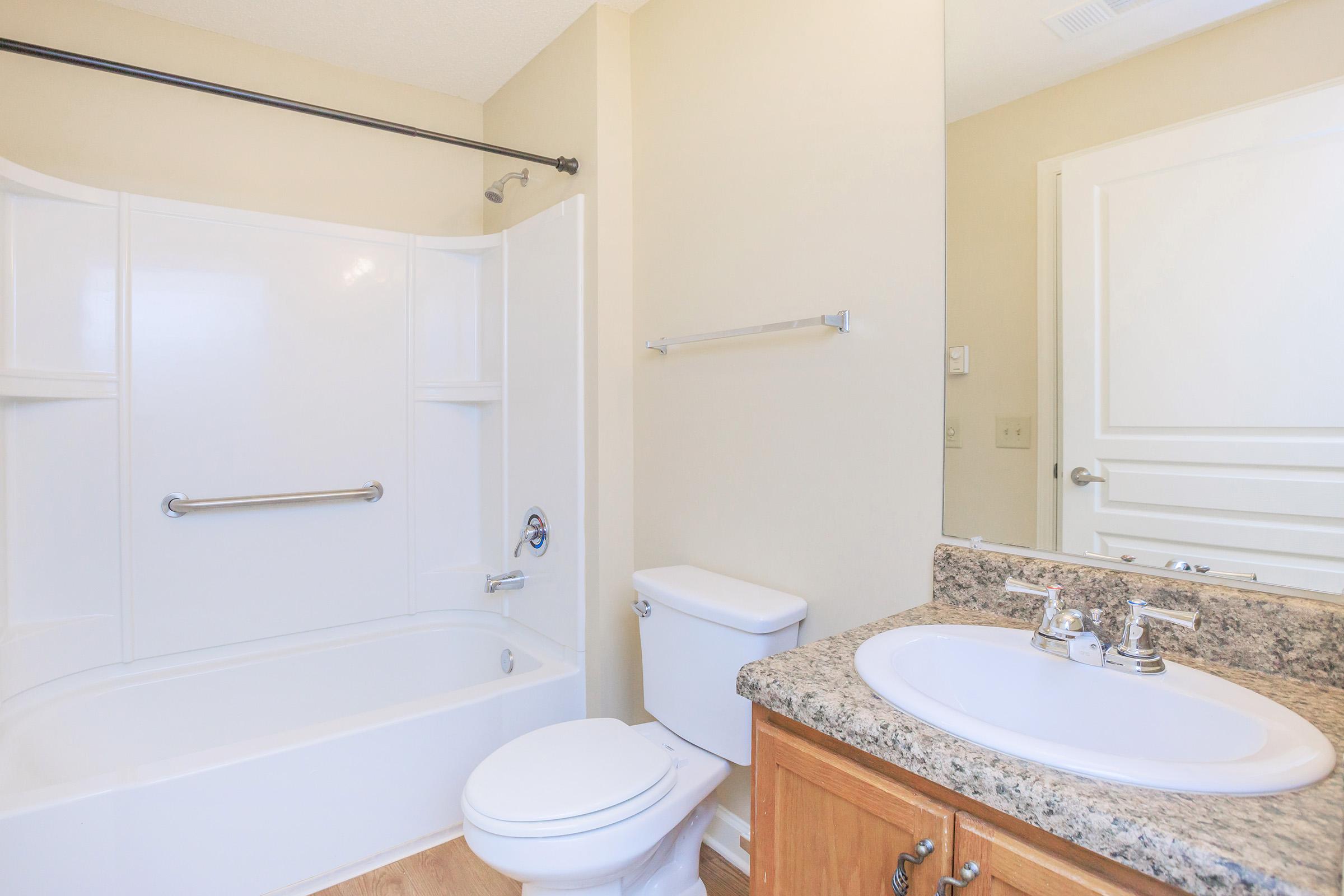 A clean, modern bathroom featuring a bathtub with a shower, a toilet, and a vanity with a sink and granite countertop. The walls are painted in a light color, and there is a simple towel bar and a mirror above the sink. The flooring is a light wood laminate.