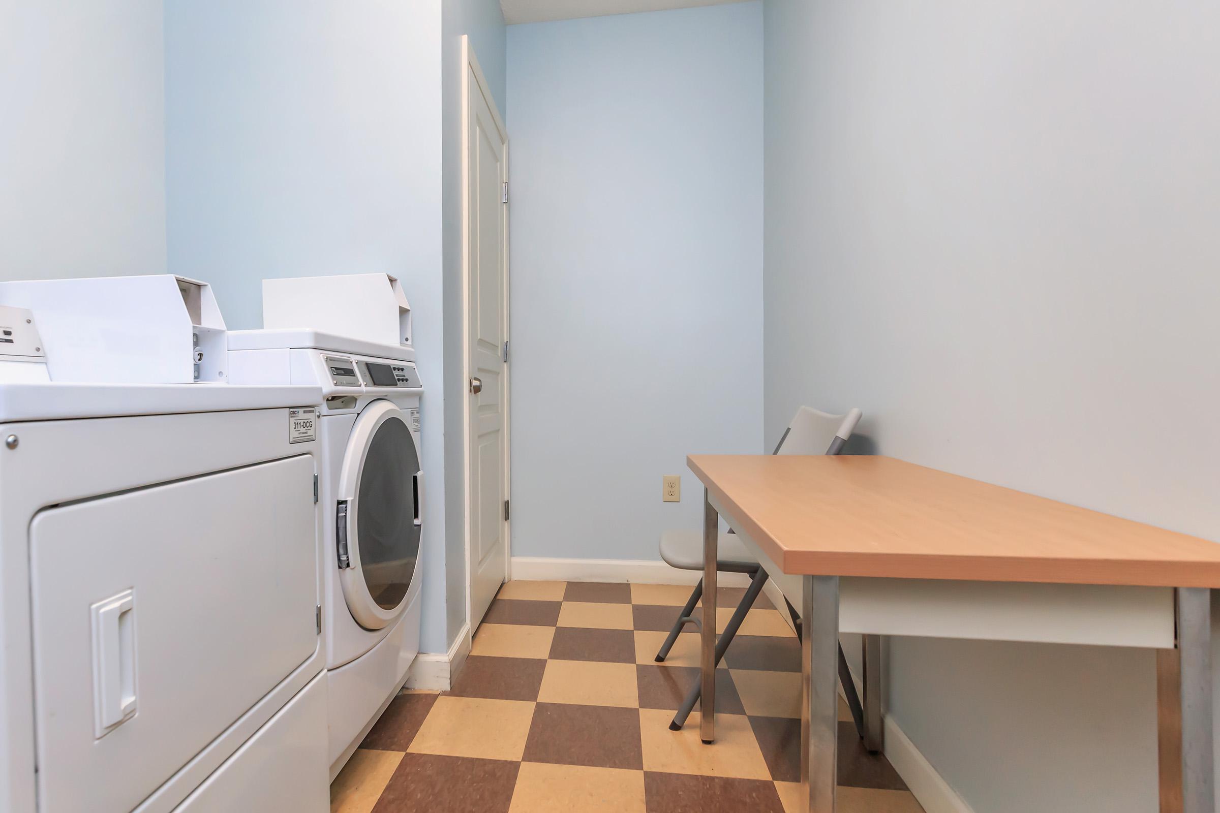 A clean, well-lit laundry room featuring a washer and dryer on the left, a folding table against the wall, and a folding chair nearby. The walls are painted light blue, and the floor has a checkered pattern in brown and beige. There's a door leading to another area.