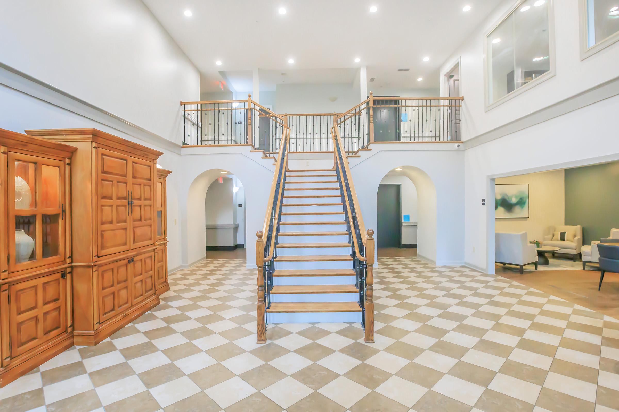 A spacious foyer featuring a central staircase with wooden handrails and a tiled floor in a diamond pattern. To the left, there's a wooden cabinet, and the space is illuminated by natural light from large windows. The walls are painted in neutral colors, creating a welcoming atmosphere.