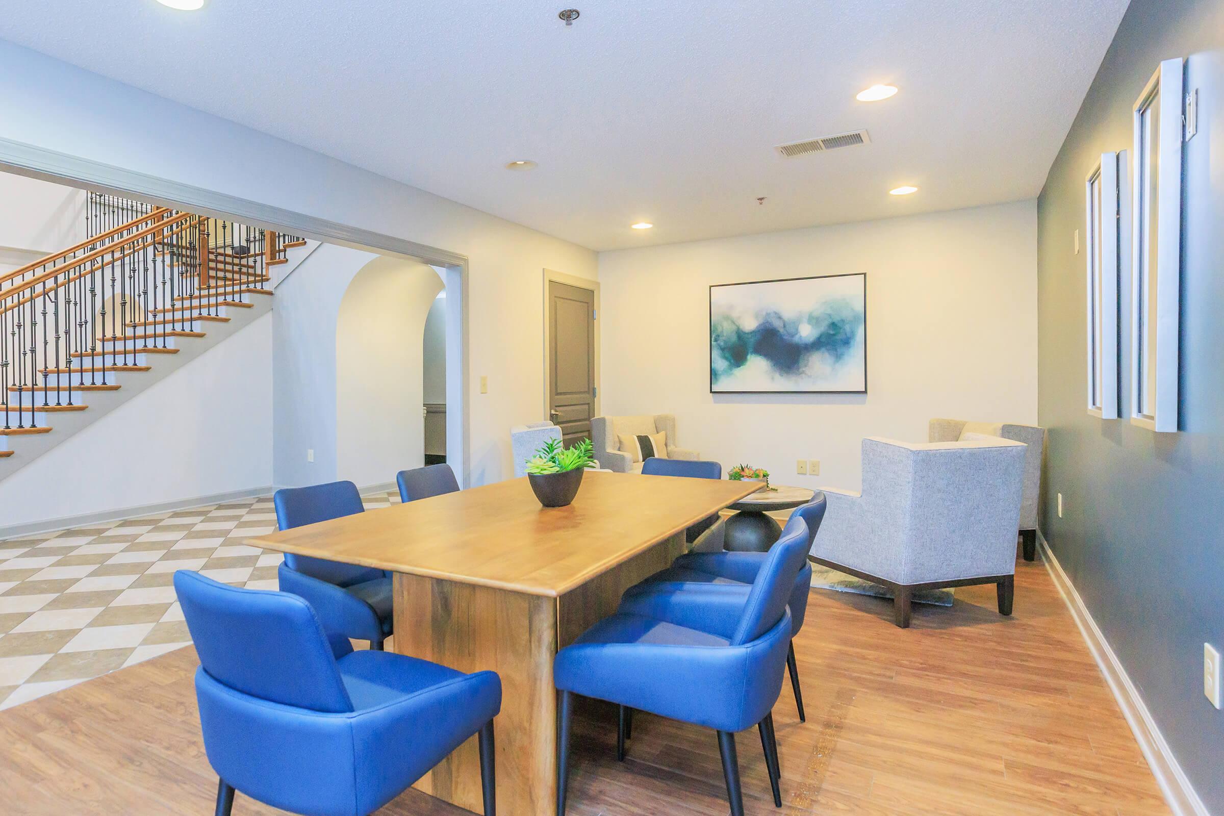 A spacious, modern living area featuring a wooden dining table surrounded by blue chairs. To one side, there are two light gray armchairs and a small potted plant on the table. The walls are painted in soft colors, and there is stylish artwork on the wall. A staircase is visible in the background.
