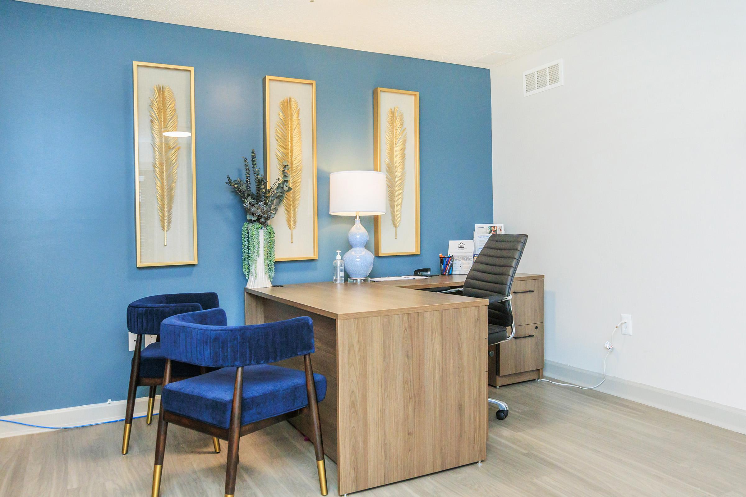 A modern office space featuring a wooden desk with a sleek black chair, two blue velvet chairs, three framed wall decorations with golden feathers, a lamp, and a vase with greenery. The walls are painted blue and white, creating a stylish and inviting atmosphere.