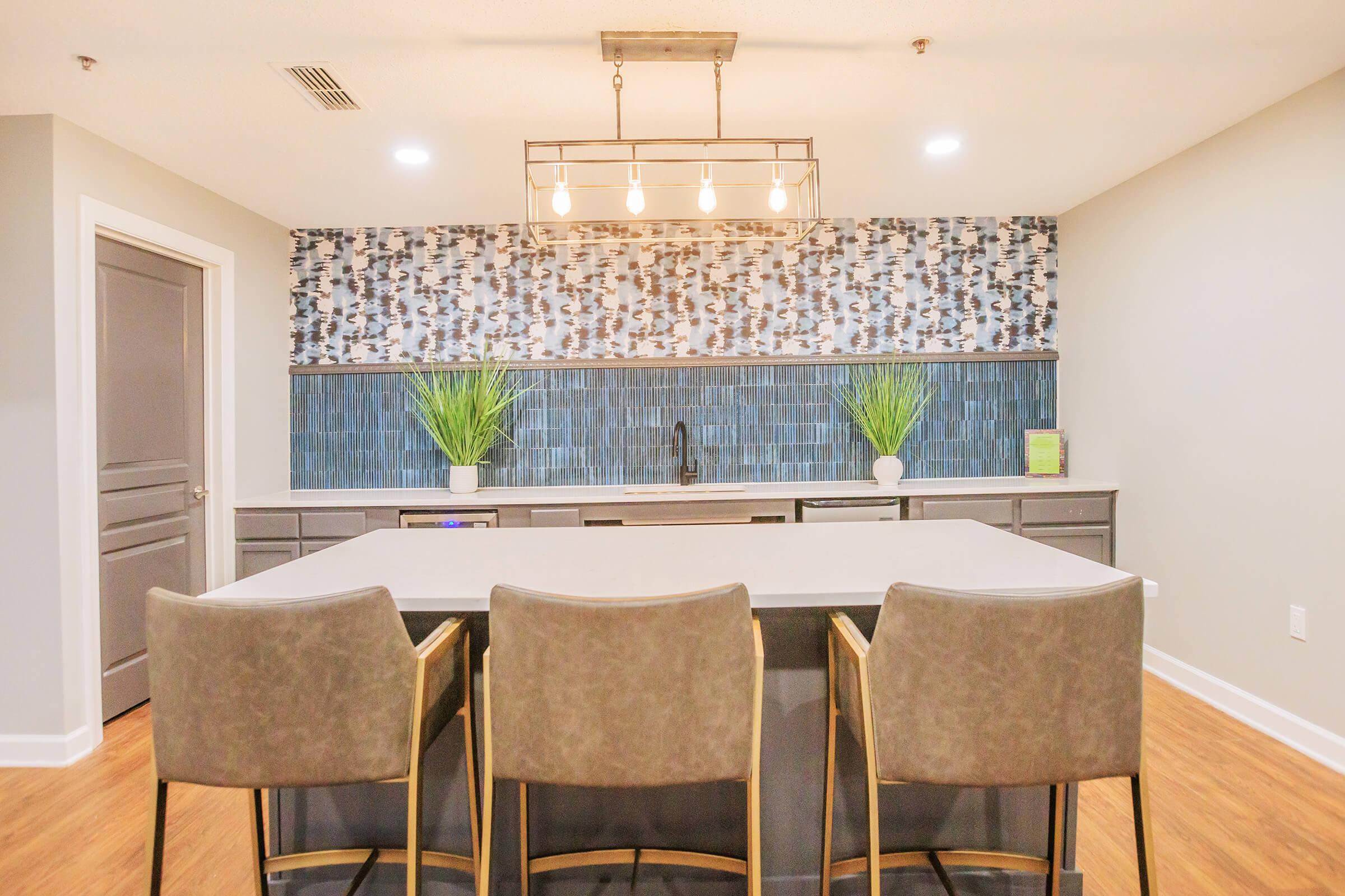 Modern kitchen with a large island featuring three cushioned bar stools. The walls are decorated with a geometric patterned backsplash and a textured tile installation. Two green plants are placed on the counter, and the lighting includes a stylish pendant fixture. The space has a contemporary design and warm wood flooring.
