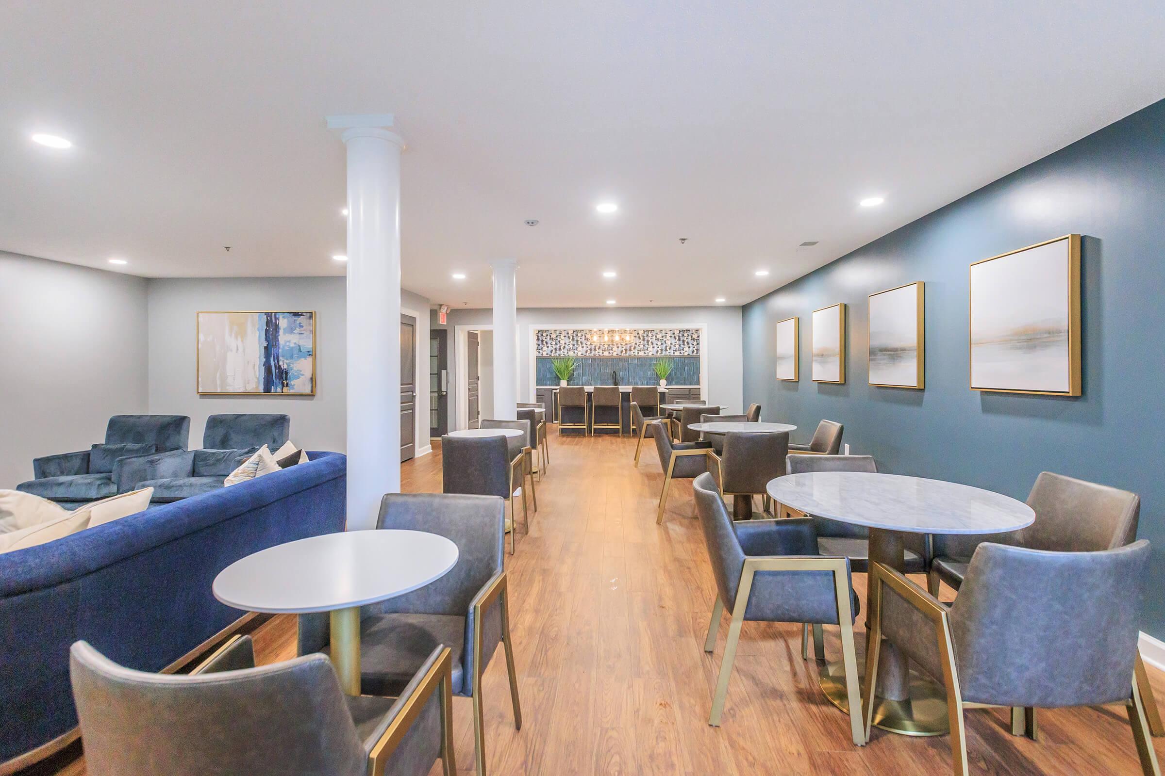 A modern lounge area featuring comfortable seating with a blue sofa, several gray chairs, and marble-topped tables. The walls are painted blue, adorned with framed artwork. A bar area is visible in the background, and the space is well-lit with soft lighting, creating an inviting atmosphere for relaxation or gatherings.