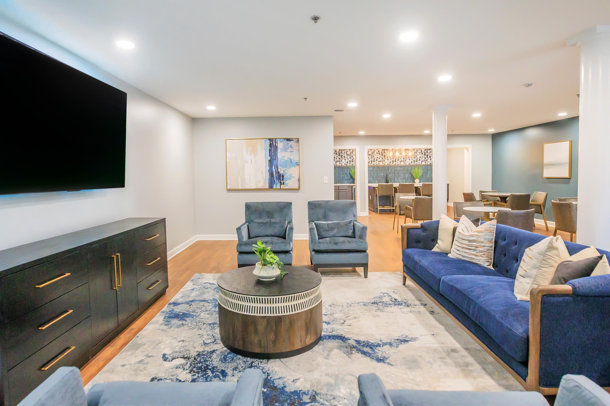A modern living space featuring a seating area with blue upholstered chairs and a matching sofa, a round coffee table with a decorative plant, and a large TV on the wall. The walls are light-colored, with an abstract art piece and a dining area visible in the background. Bright lighting enhances the open and inviting atmosphere.