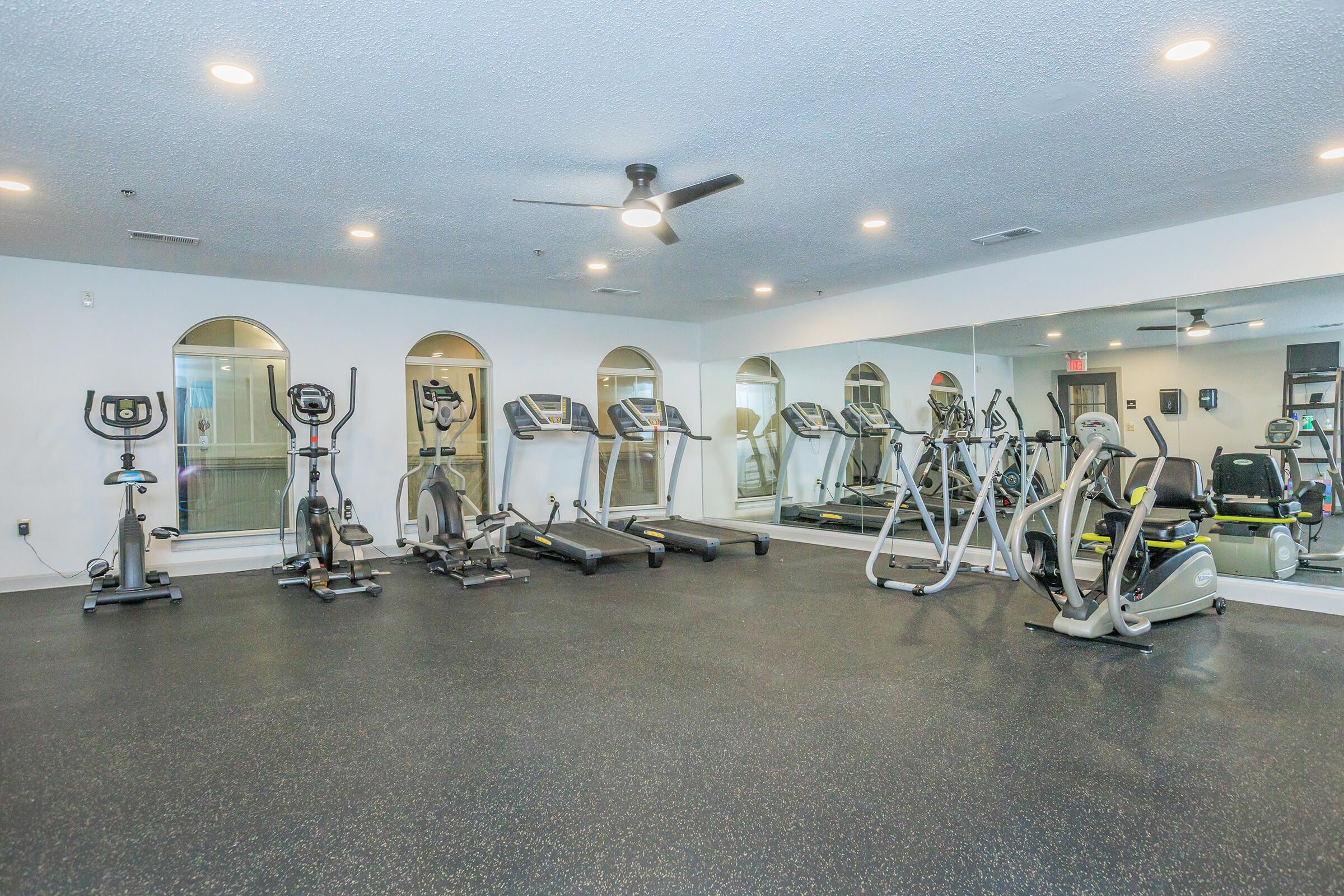 A modern gym interior featuring multiple treadmills, elliptical machines, and stationary bikes, all arranged neatly on a rubber floor. The space is well-lit with overhead lighting and has large mirrors along the walls, creating a spacious and inviting workout environment.